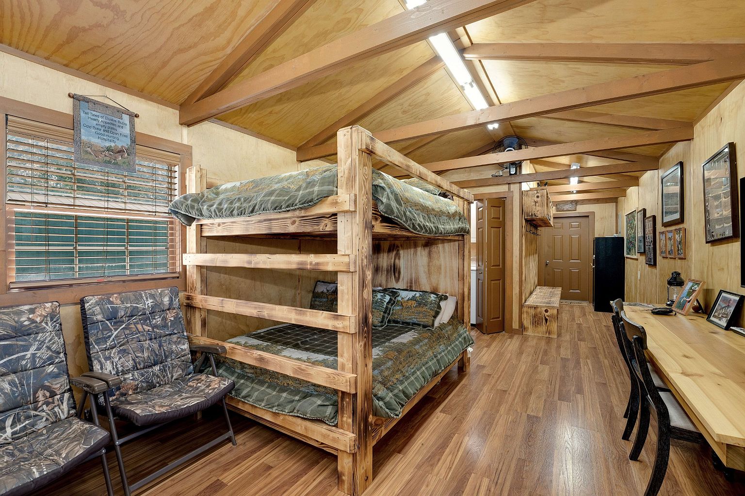 This is a rustic bedroom featuring a wooden bunk bed with patterned bedding, complemented by two camouflage-patterned chairs. The room has wood-paneled walls and hardwood floors, creating a cozy, cabin-like atmosphere. A desk and chair set are visible in the background, suggesting a functional space for work or study.
