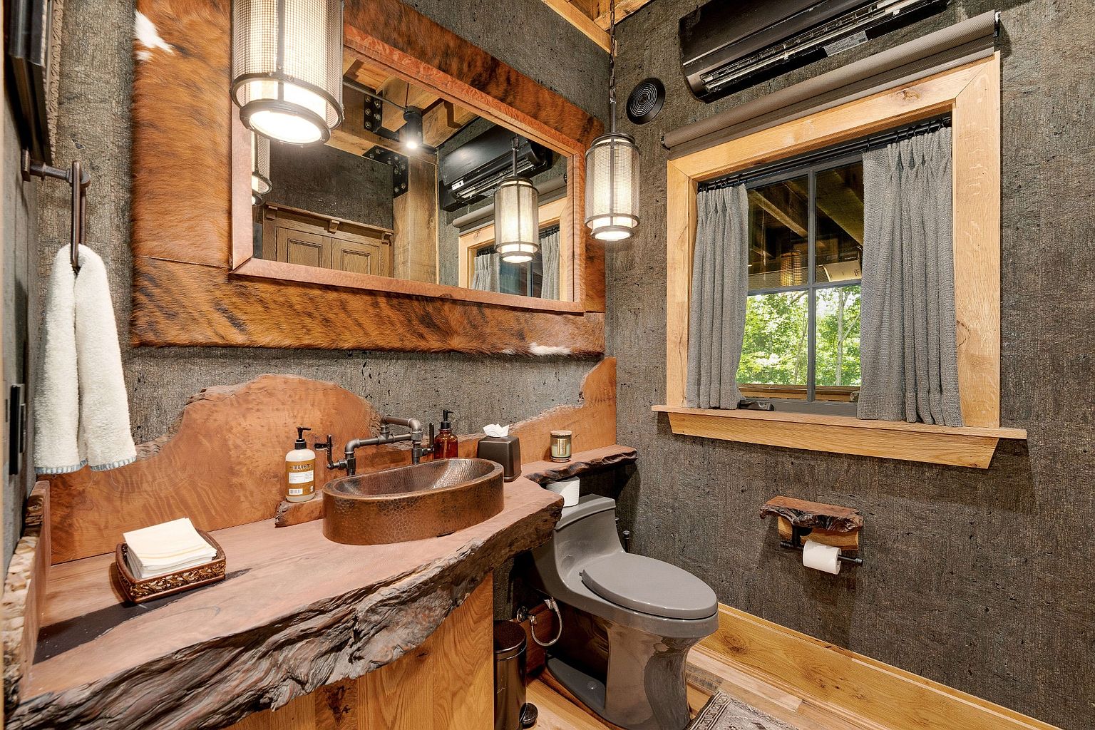This is a rustic bathroom featuring a unique live-edge wood countertop with a copper vessel sink and industrial-style faucet. The walls are textured in a dark gray, complemented by a wood-framed mirror and pendant lighting. A modern toilet and window with gray curtains add to the room's blend of natural and contemporary elements.
