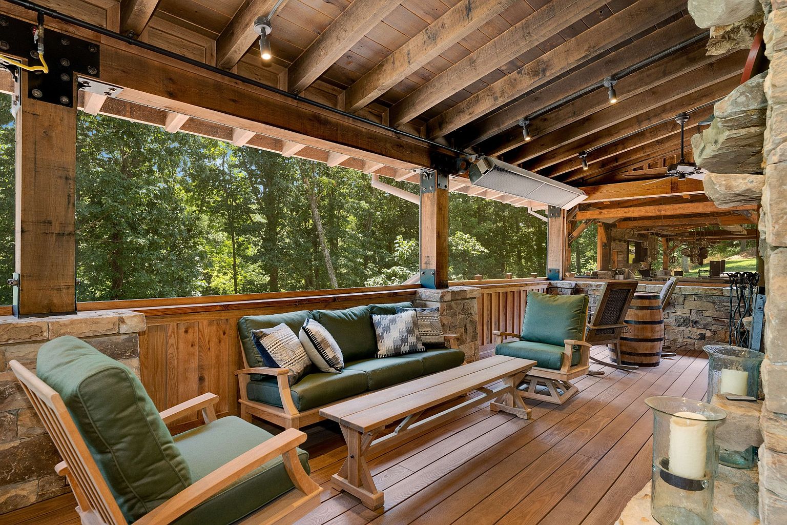 This image showcases a beautifully designed outdoor patio or deck area. The space features comfortable seating arrangements with green cushioned chairs and a sofa, complemented by a wooden coffee table. The wooden deck flooring and ceiling, along with stone accents, create a warm and inviting atmosphere, perfect for relaxation and entertaining.