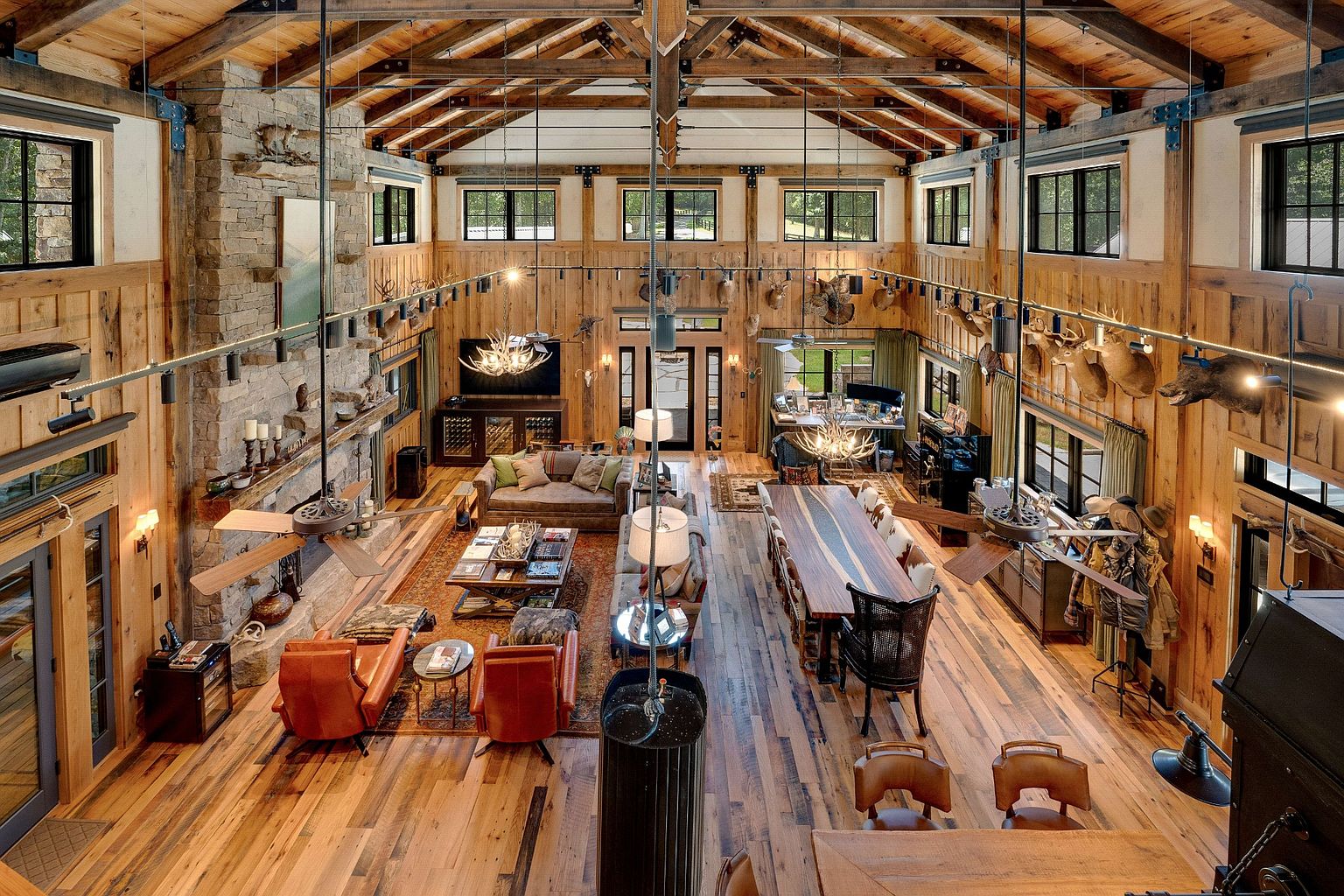 This is a grand living room with a rustic, lodge-style design. The room features high ceilings with exposed wooden beams, wood paneled walls adorned with mounted animal heads, and a stone fireplace. The space is furnished with leather seating, a large dining table, and various decorative elements, creating a warm and inviting atmosphere.