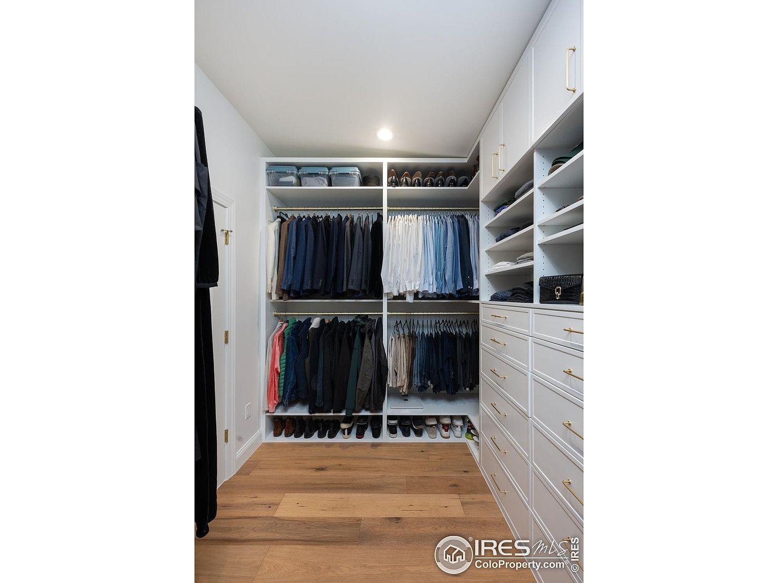This is a well-organized walk-in closet featuring custom white cabinetry with gold hardware. The closet includes shelving, hanging rods for clothing, drawers, and shoe storage. The hardwood flooring adds a touch of warmth to the space, creating a luxurious and functional area for clothing and accessories.
