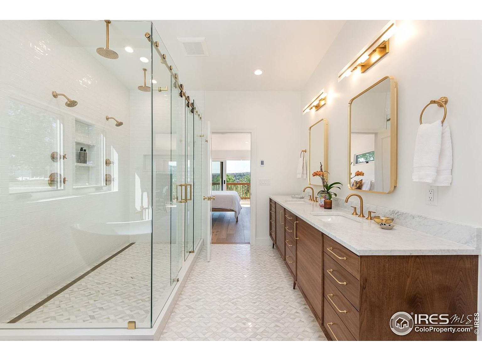 This is a bright and modern primary bathroom featuring a large glass-enclosed shower with multiple shower heads and white subway tile. The double vanity has a marble countertop, wood cabinets with gold hardware, and gold-framed mirrors. The flooring is a patterned tile, and the overall aesthetic is luxurious and spa-like.