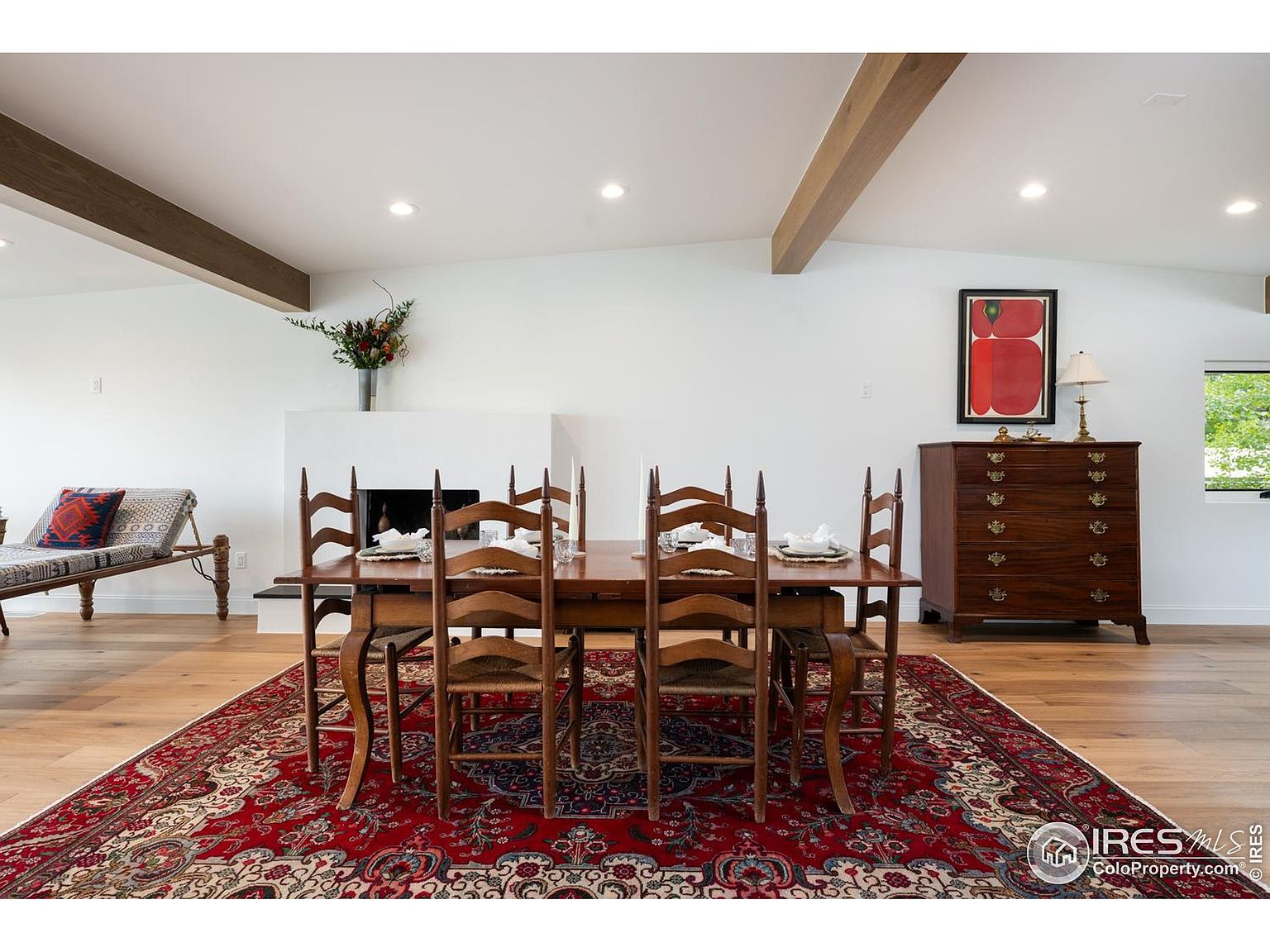 This is an interior shot of a dining room featuring a long wooden table with six chairs set on a red patterned rug. The room has white walls, wooden beams on the ceiling, and a fireplace. A wooden chest of drawers sits against the wall with a lamp and artwork above it, creating a warm and inviting atmosphere.