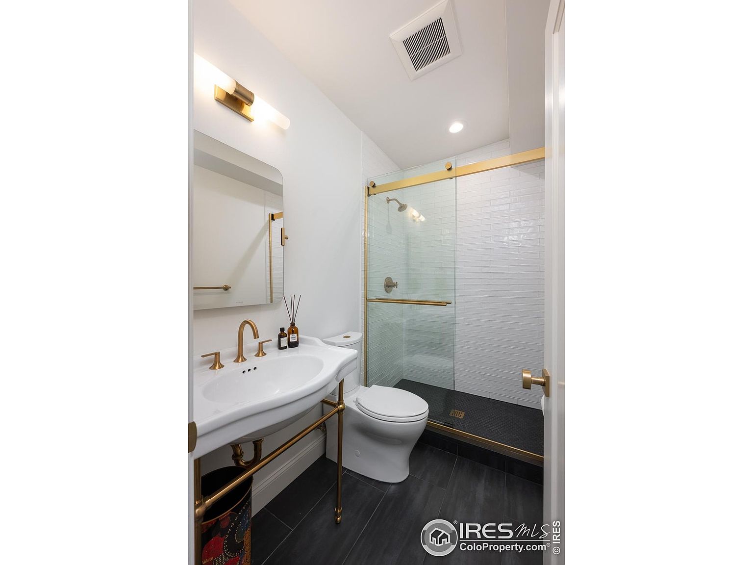 This is a well-lit bathroom featuring a white sink with gold fixtures, a matching mirror, and a toilet. The shower has a glass door with gold trim and white tiled walls. The floor is dark gray, creating a modern and clean aesthetic.