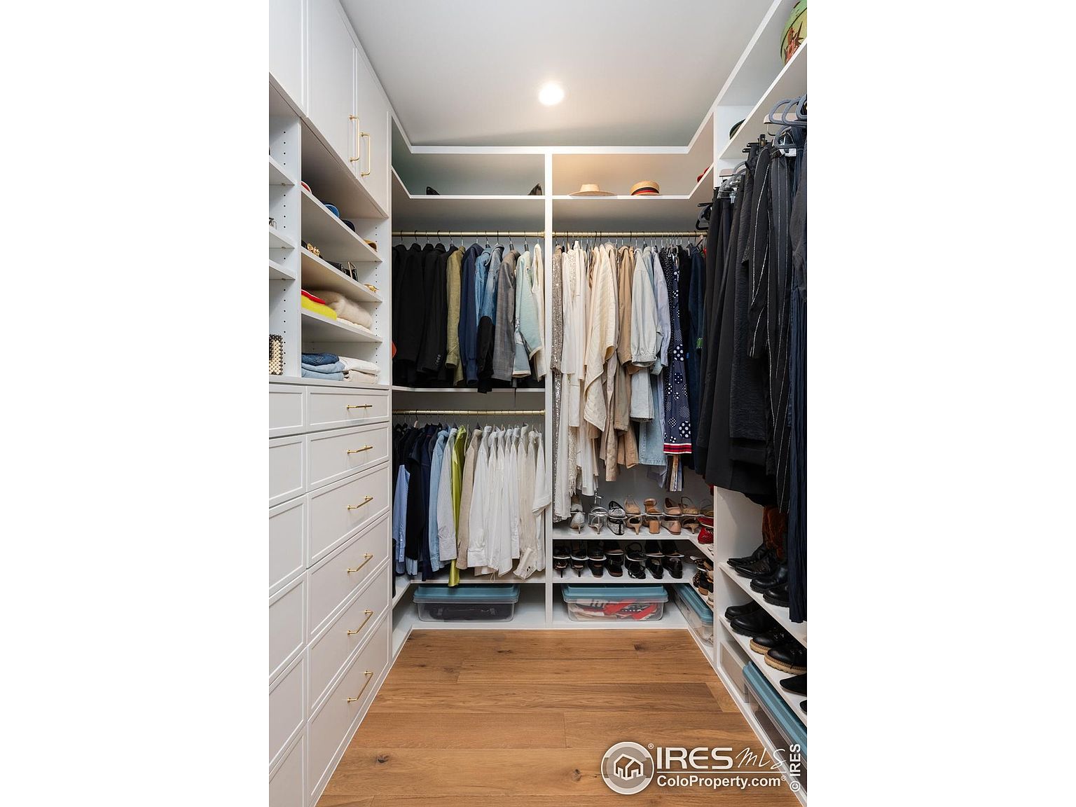 This is a well-organized walk-in closet with white cabinetry and drawers, gold hardware, and wooden flooring. The closet features hanging rods for clothes, shelves for shoes and folded items, and storage bins. The space is brightly lit and appears spacious, offering ample storage for clothing and accessories.