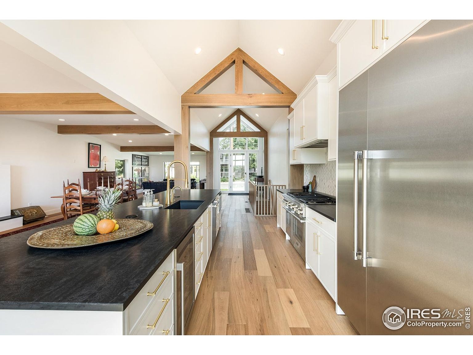 This is a bright, open kitchen with a modern farmhouse aesthetic. It features a large island with a dark countertop, white cabinetry with gold hardware, and stainless steel appliances. The kitchen has a high vaulted ceiling with exposed wooden beams, and it opens into a dining and living area, creating a spacious and inviting atmosphere.