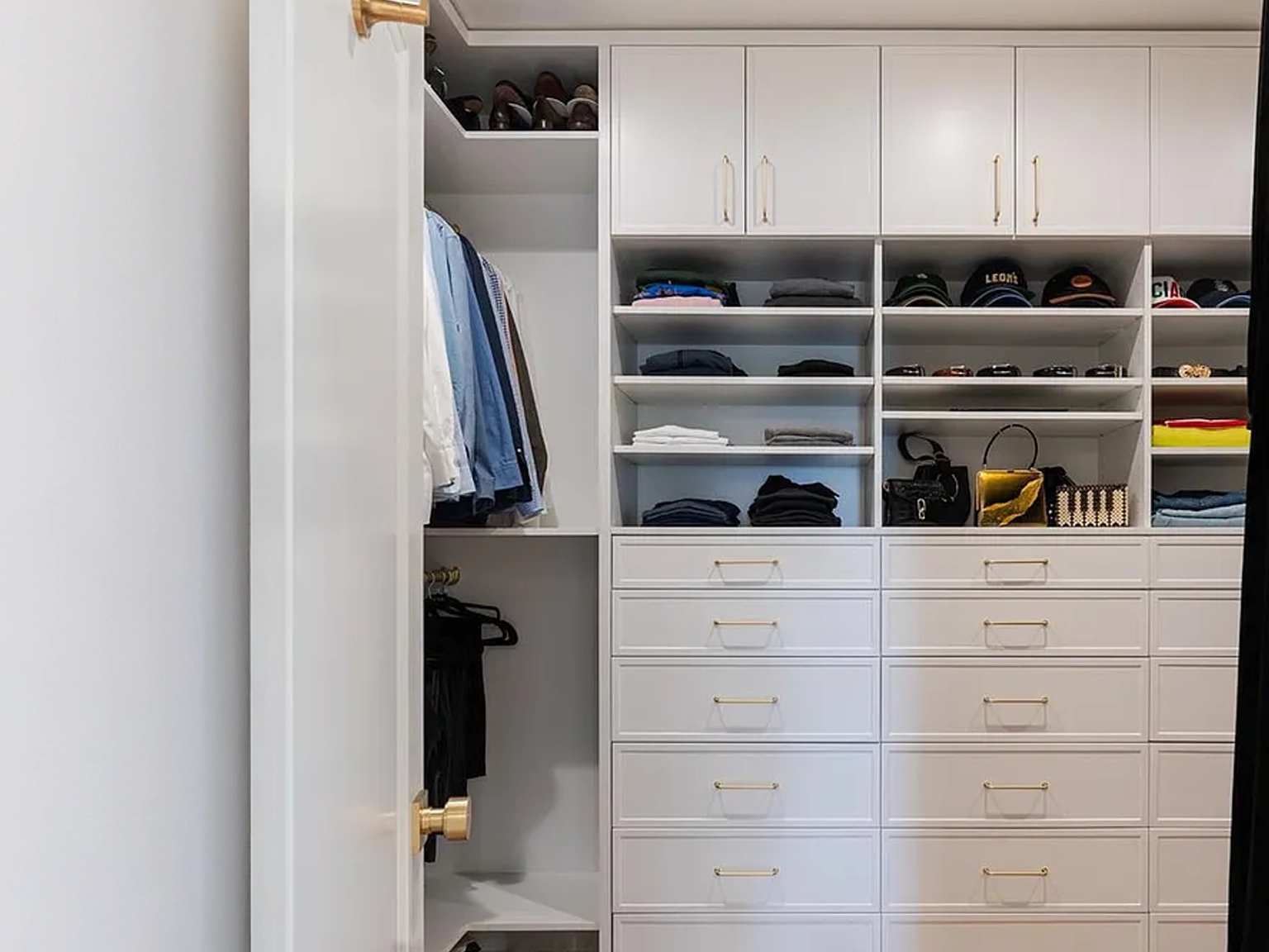 This is an interior shot of a well-organized walk-in closet. The closet features white cabinetry with gold hardware, including various shelves, drawers, and rods for hanging clothes. The design offers ample storage space in a stylish and functional manner.