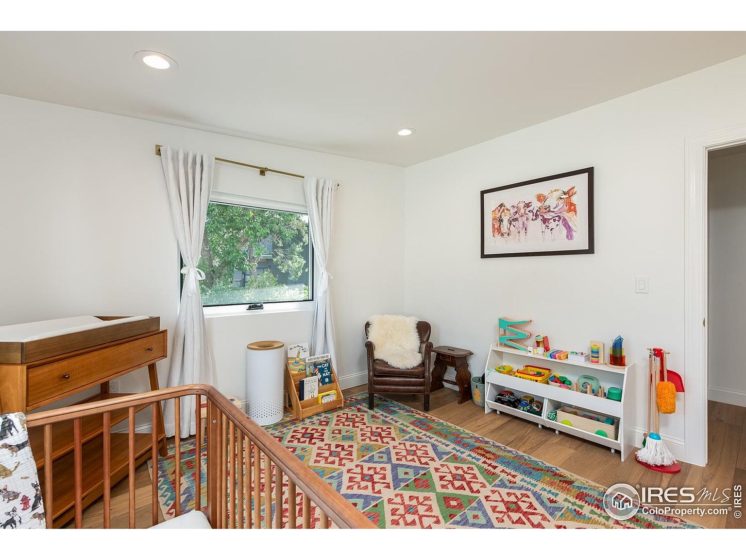 This is an interior shot of a child's bedroom, showcasing a cozy and well-organized space. The room features a wooden crib, a changing table, a colorful patterned rug, and a toy shelf filled with various toys. A leather armchair with a sheepskin throw adds a touch of comfort, while a framed picture hangs on the wall, creating a playful and inviting atmosphere.