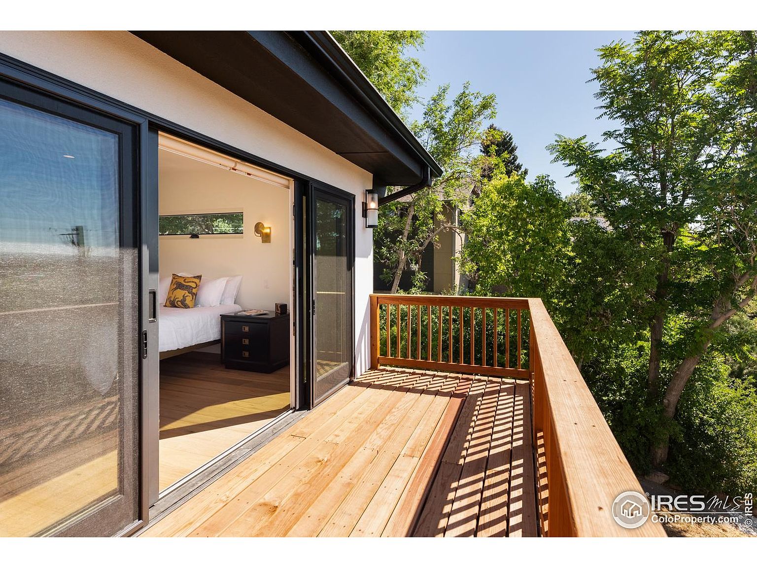 This image showcases a charming outdoor deck or balcony area, seamlessly connected to an interior bedroom through sliding glass doors. The deck features wooden flooring and a matching wooden railing, offering a warm and inviting atmosphere. Lush greenery and trees surround the deck, providing privacy and a sense of tranquility.
