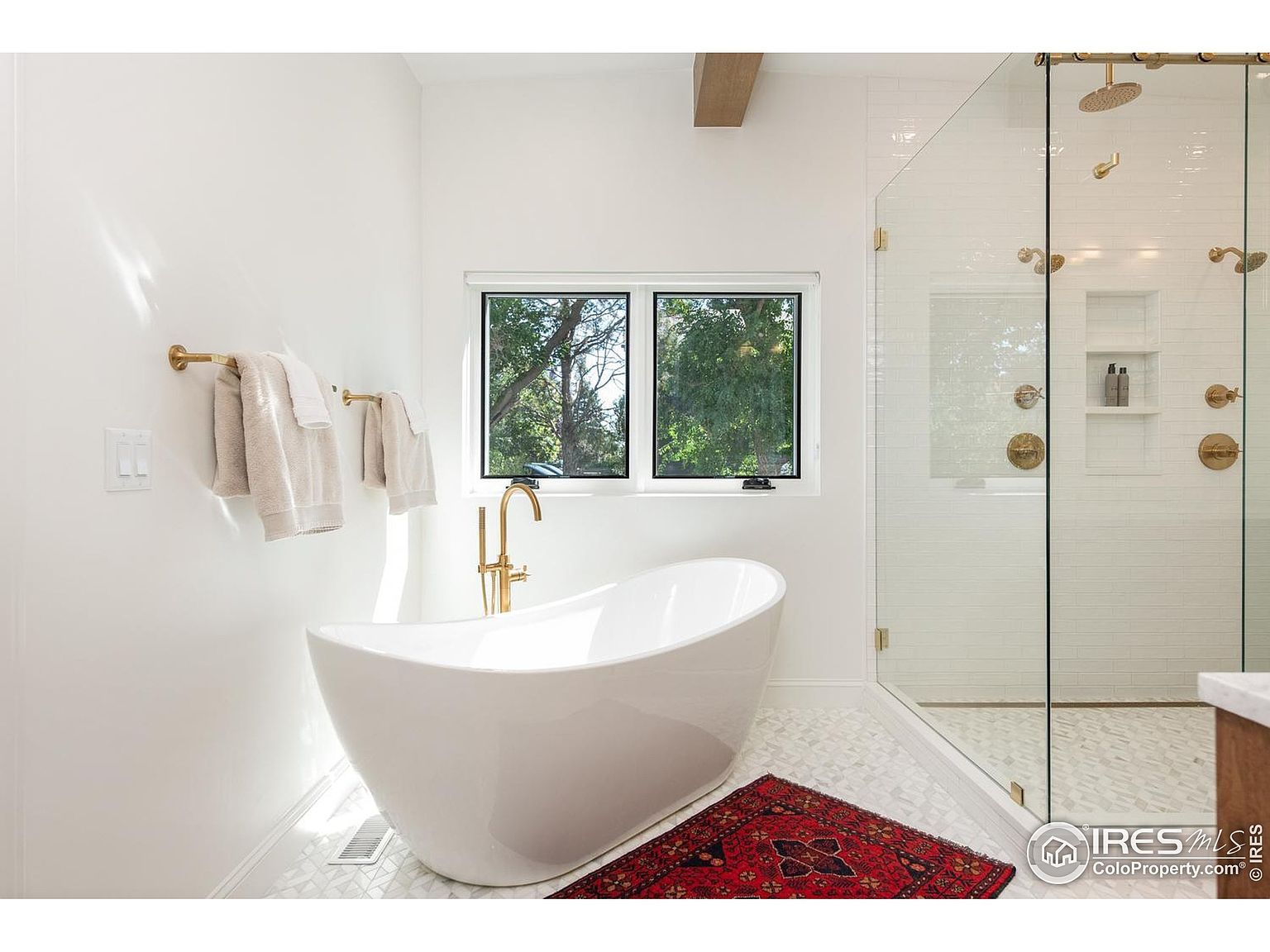 This is a bright and modern primary bathroom featuring a freestanding white bathtub with gold fixtures, a large window providing natural light, and a glass-enclosed shower with gold hardware. The floor is tiled with a geometric pattern, and a small red rug adds a pop of color. The overall impression is clean, luxurious, and well-designed.