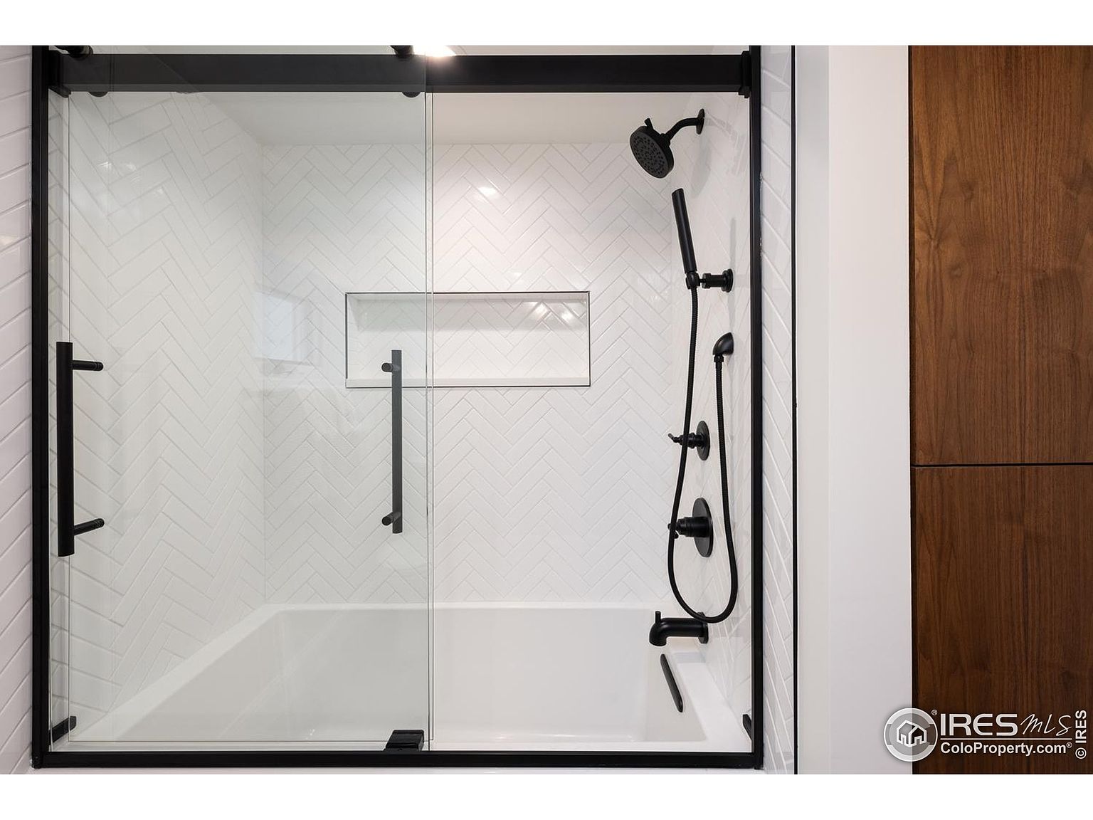 This is a well-lit bathroom featuring a modern bathtub and shower combination. The shower has a sleek, black-framed glass enclosure with a sliding door, complemented by black fixtures including the shower head and faucet. The walls are adorned with white herringbone-patterned tiles, and a built-in niche provides storage space.