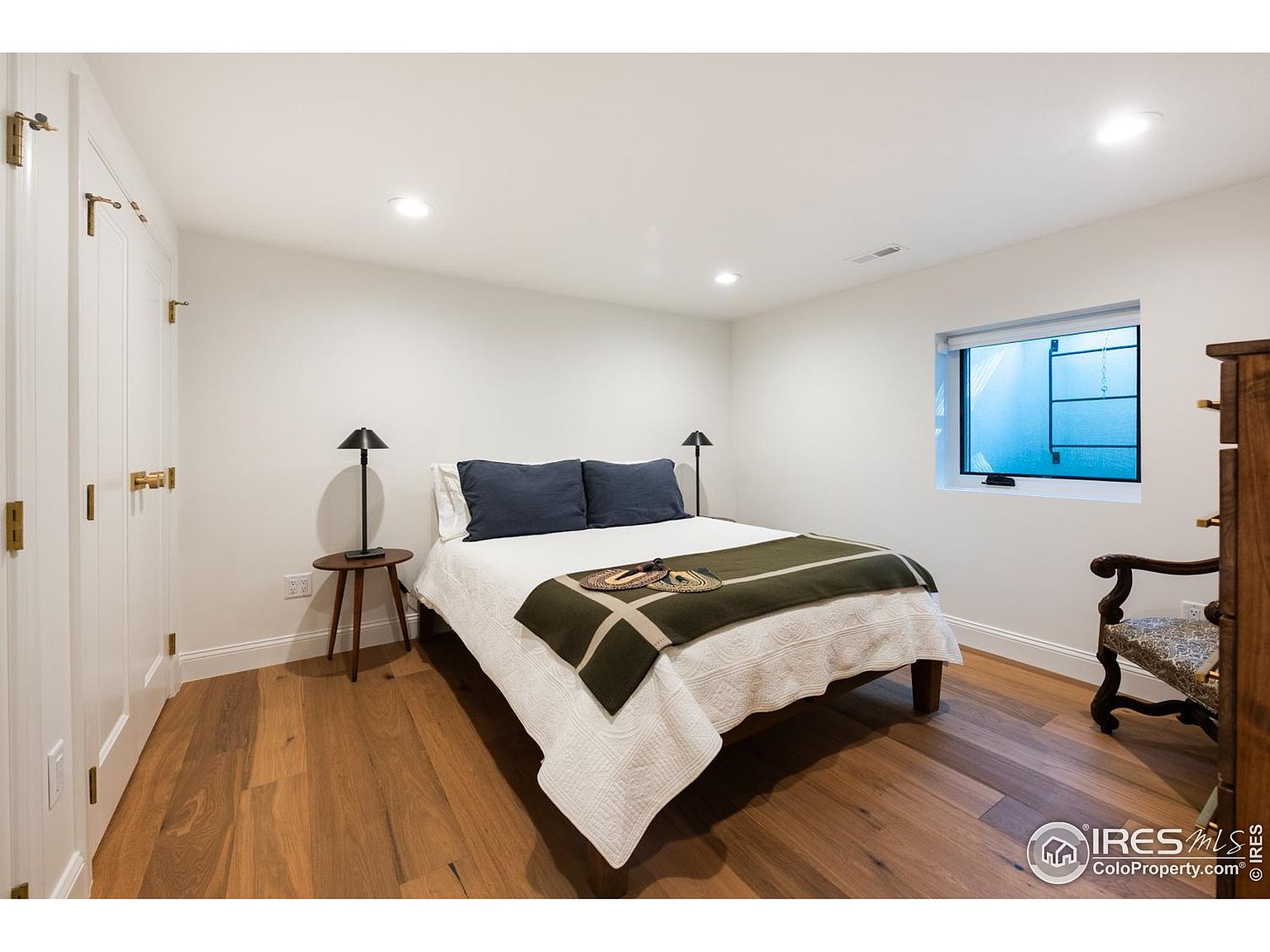 This is a cozy bedroom featuring hardwood floors, white walls, and a bed with blue pillows and a green blanket. A small side table with a lamp sits next to the bed, and a window provides natural light. The room has a simple and clean design, creating a comfortable and inviting atmosphere.
