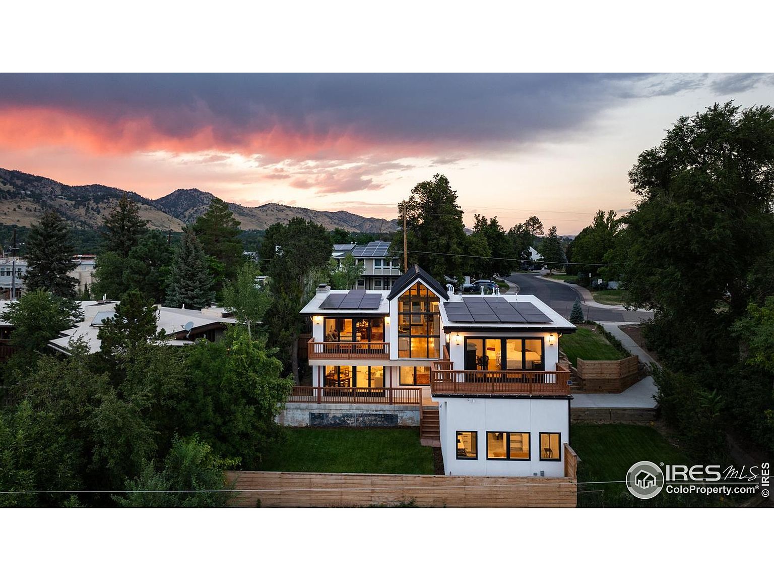 This aerial view showcases a modern two-story home with a combination of white siding and wood accents. Solar panels are visible on the roof, and multiple balconies offer outdoor living spaces. The property is surrounded by lush greenery and mature trees, with mountains visible in the background, creating a serene and private setting.