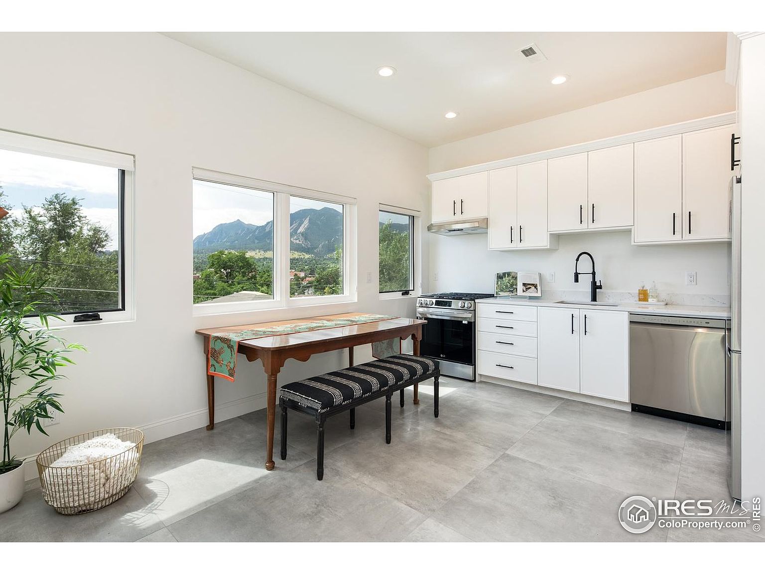 This is a bright and modern kitchen featuring white cabinets, stainless steel appliances, and a sleek black faucet. A wooden dining table with a striped bench sits adjacent to the kitchen area, offering a casual dining space with a view of the mountains through the large windows. The gray tile flooring and minimalist decor contribute to a clean and contemporary aesthetic.