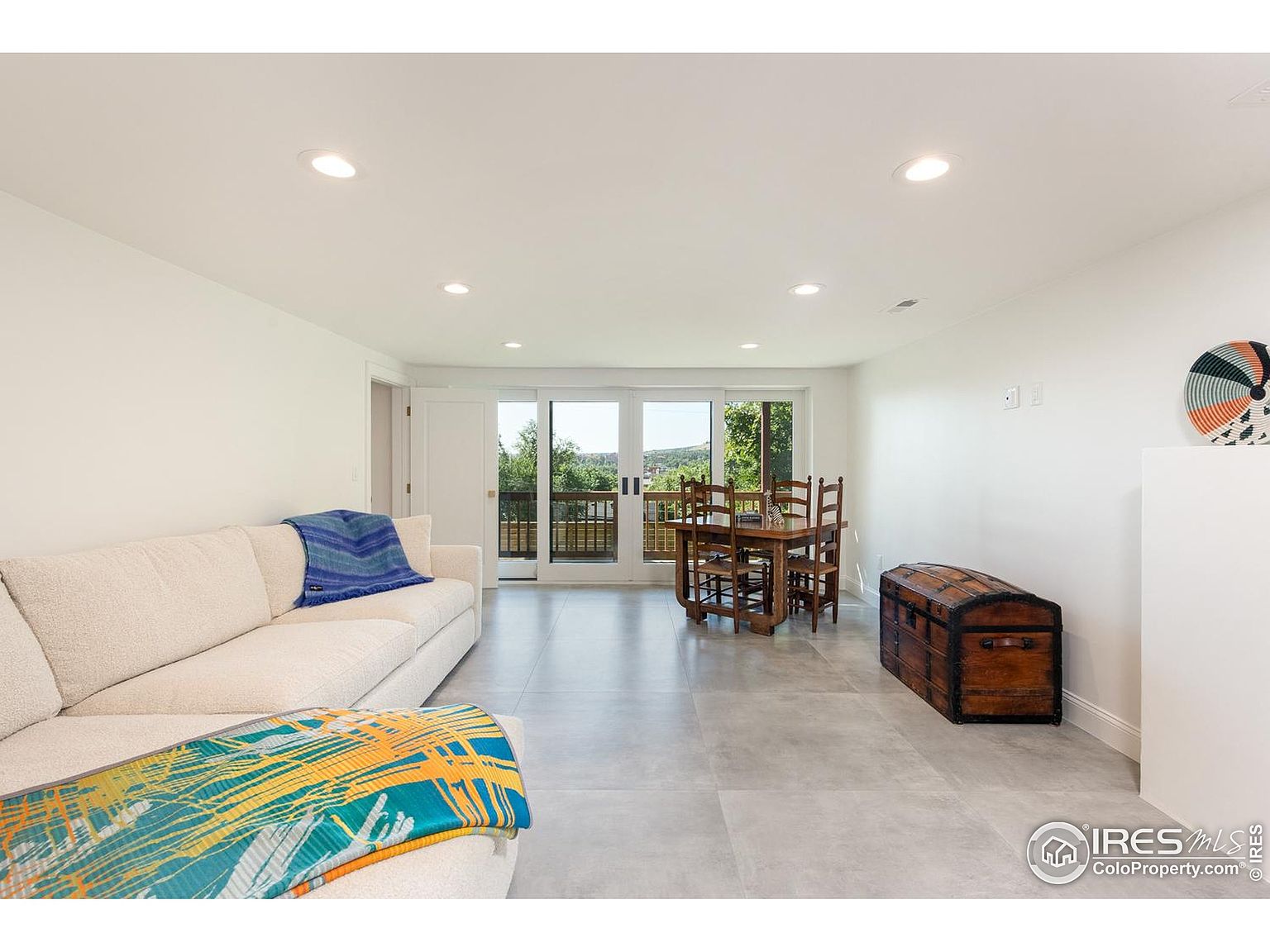 This is an interior shot of a living room area, featuring a neutral-toned sofa with blue and patterned throws, a wooden dining table with chairs, and a decorative wooden chest. The room has light gray tile flooring, white walls, and recessed lighting, creating a bright and airy atmosphere. Sliding glass doors lead to an outdoor balcony, enhancing the sense of space and connection to the outdoors.