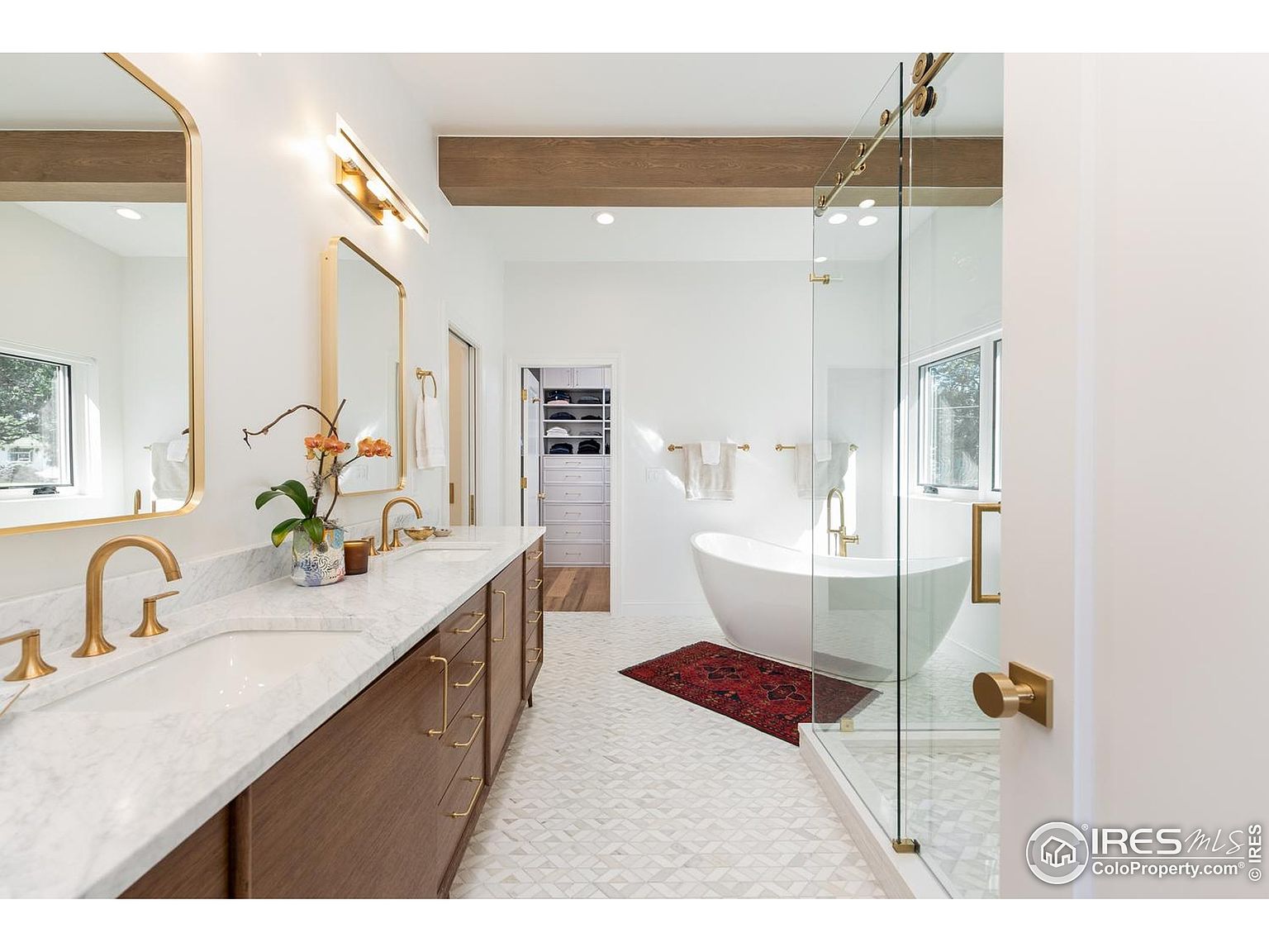 This is a bright and modern primary bathroom featuring a double vanity with marble countertops and wood cabinetry. A freestanding bathtub sits near a glass-enclosed shower, and the space is accented with gold fixtures and hardware. The geometric tile flooring and exposed wood beam add a touch of contemporary elegance.