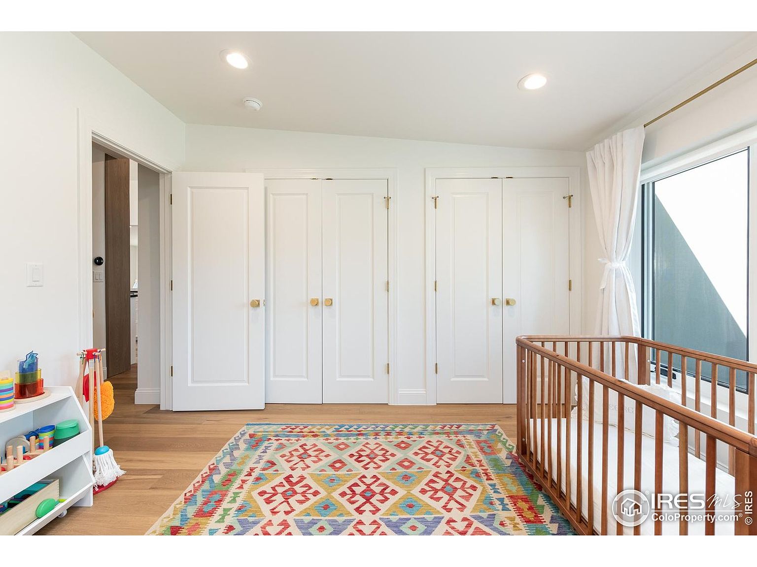 This is a bright and airy bedroom featuring three white closet doors with gold hardware, a colorful patterned rug, and a wooden crib. Natural light floods the room through a large window with white curtains. The hardwood flooring adds warmth to the space, creating a cozy and inviting atmosphere.