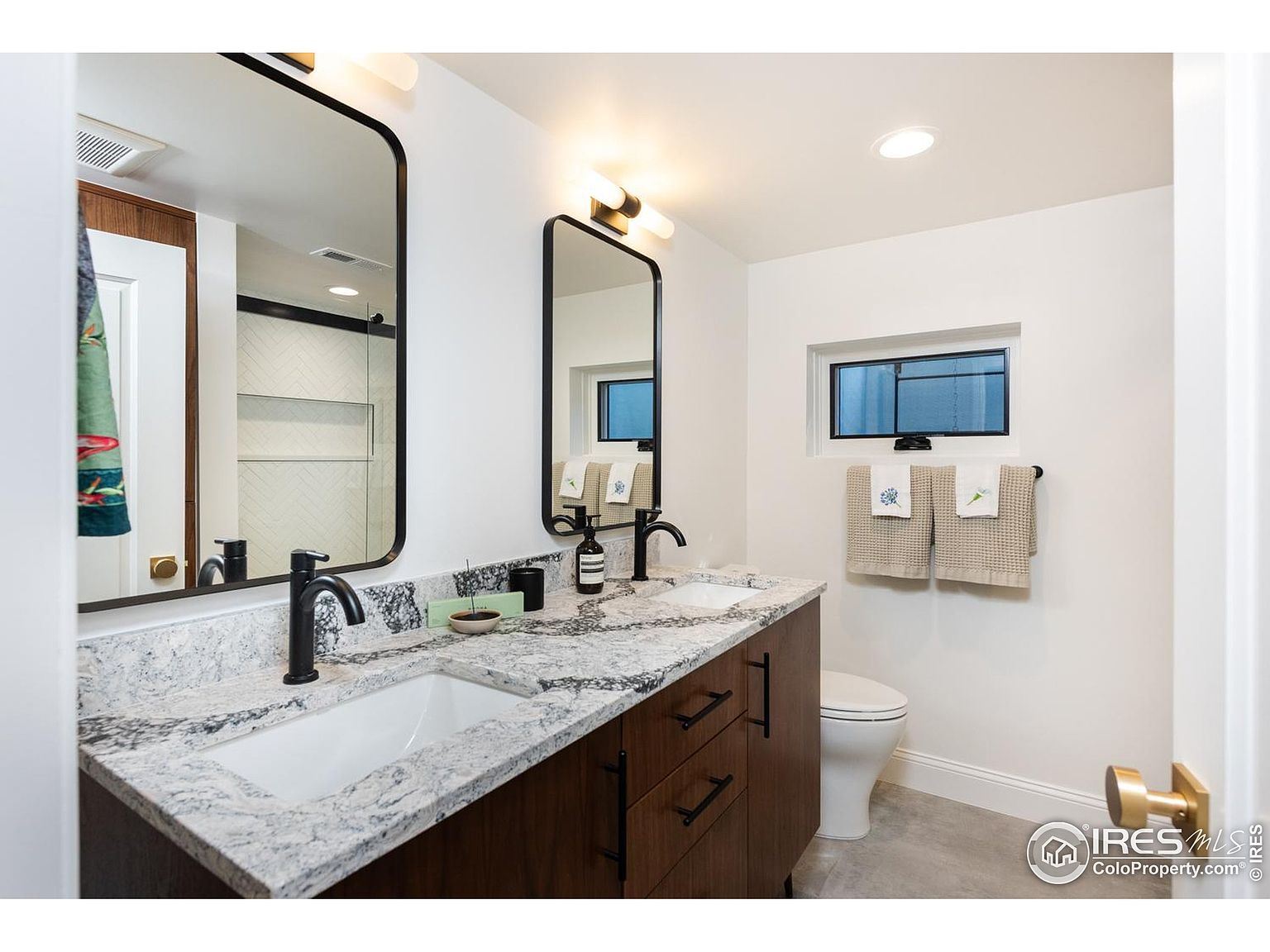 This is a well-lit bathroom featuring a double vanity with a granite countertop and dark wood cabinetry. Two black-framed mirrors hang above the sinks, complemented by modern light fixtures. A toilet is visible to the right, and a small window provides natural light, creating a clean and contemporary space.