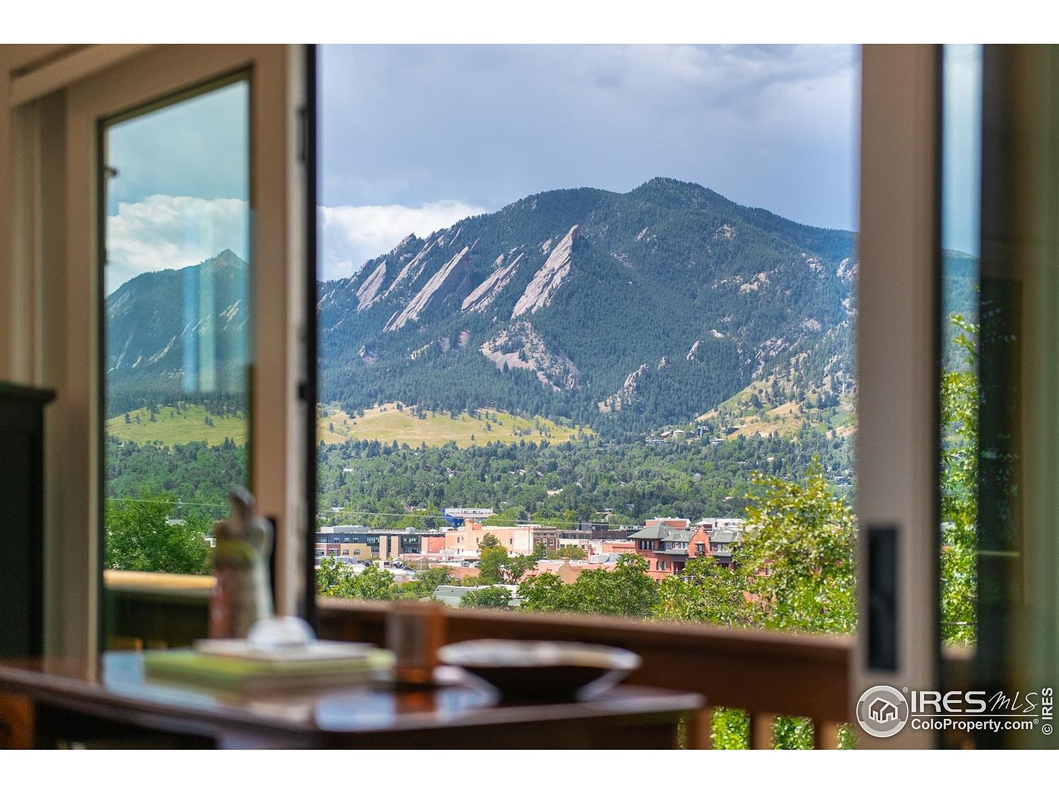 This image showcases a view from inside a living room, looking out through a large open window or doorway. The focus is on the scenic mountain vista, with a glimpse of the town below. The interior elements, such as the table and decor, are slightly blurred, emphasizing the view as the primary feature.