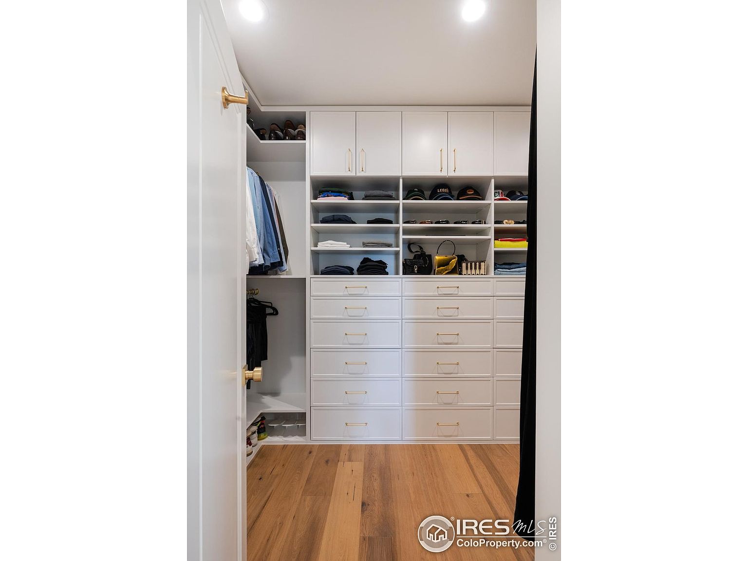 This image showcases a well-organized walk-in closet with custom white cabinetry and gold hardware. The closet features ample shelving, drawers, and hanging space, providing plenty of storage. The hardwood flooring adds a touch of warmth to the space, creating a luxurious and functional area.
