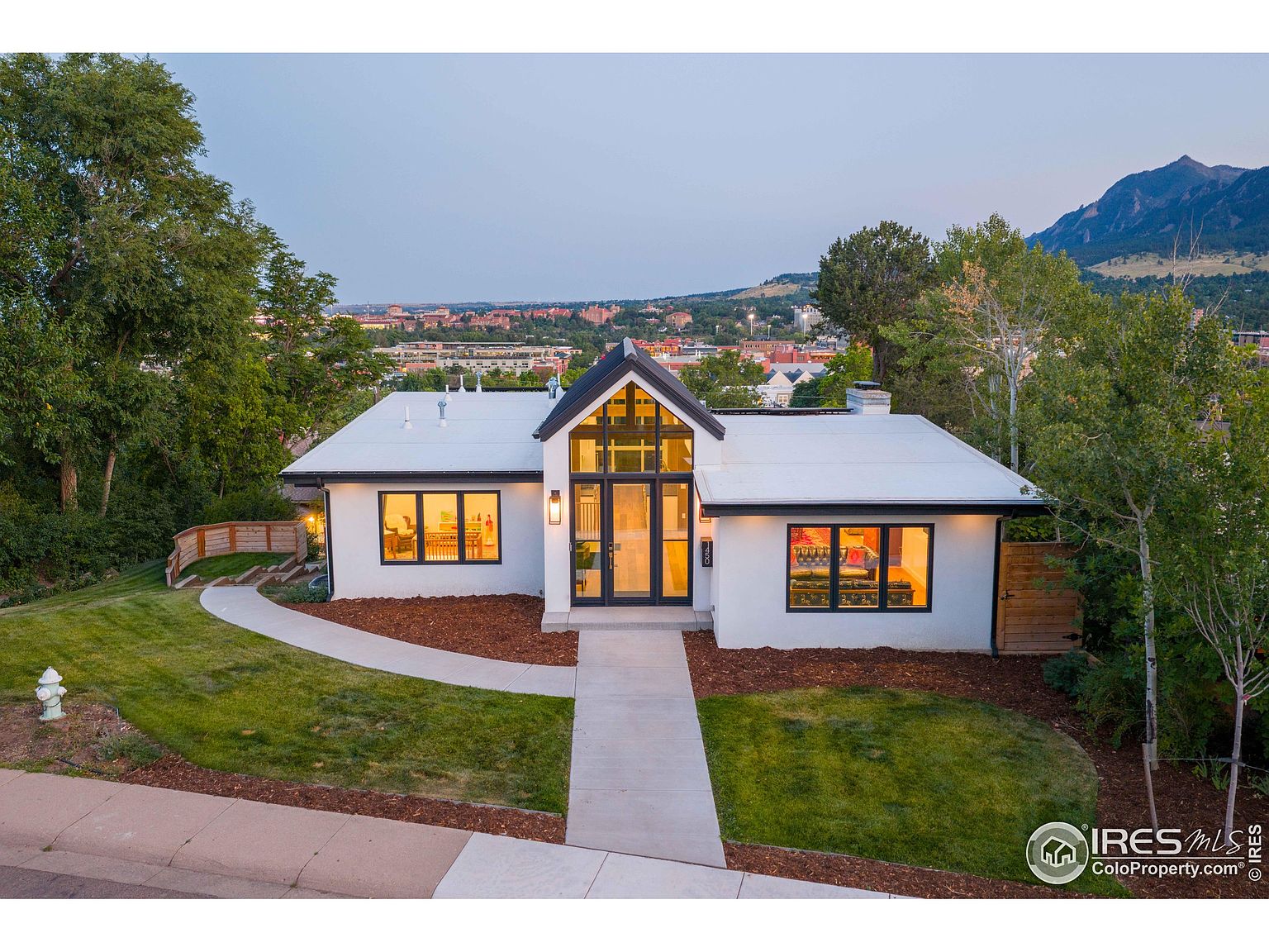 This is a front exterior view of a modern home with a white facade and a striking black-framed entryway. The house features a well-manicured lawn, a concrete walkway leading to the front door, and mature trees in the surrounding landscape. The mountain range in the background adds to the property's appeal.