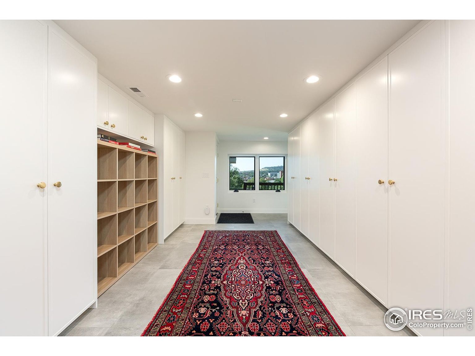 This is a well-organized walk-in closet featuring white cabinetry with gold hardware on both sides of a long hallway. A red patterned rug runs down the center of the gray tile floor, leading to a window with a view of the outdoors. A wooden shelving unit is on the left side of the closet.