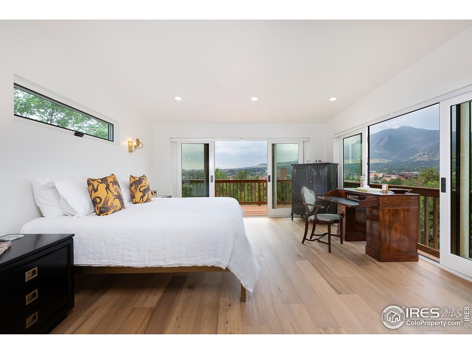This is a bright and airy primary bedroom featuring hardwood floors, white walls, and large sliding glass doors that lead to a balcony with mountain views. The room includes a bed with white linens and decorative pillows, a dark wood nightstand, a desk area with a chair, and a dark wood cabinet. The overall impression is one of tranquility and connection to nature.