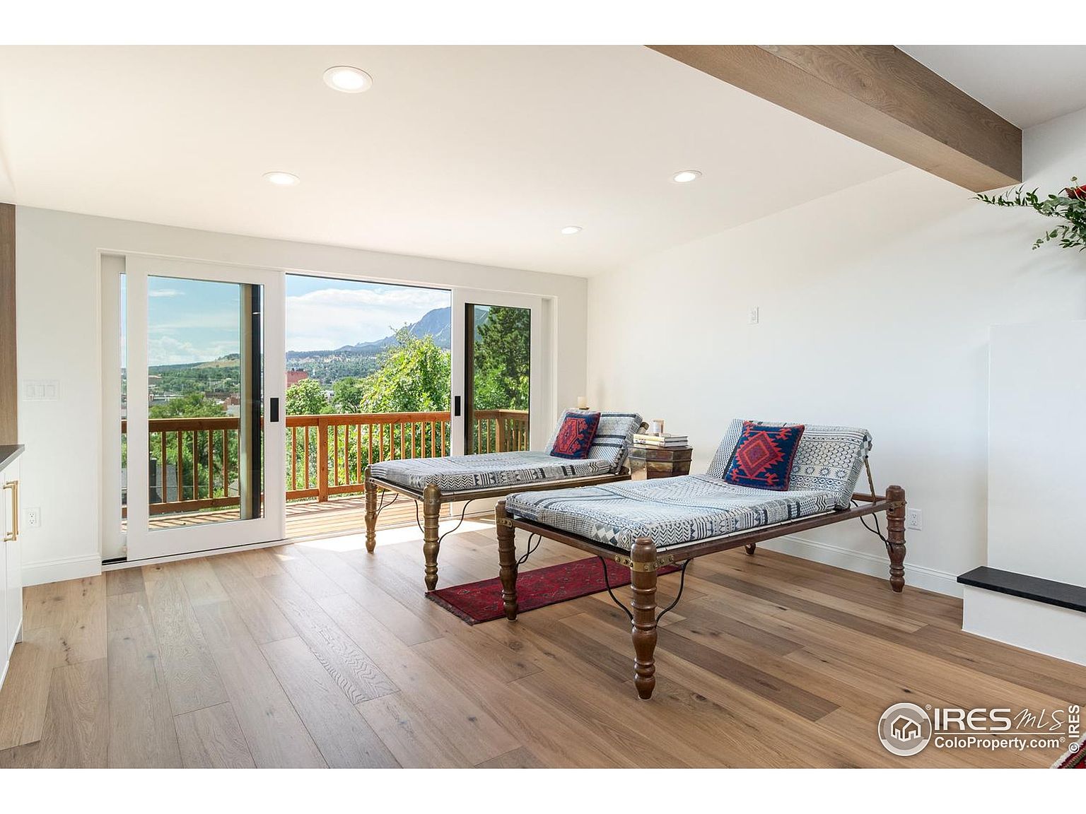 This interior shot showcases a bright living room with hardwood floors and a sliding glass door leading to a balcony with mountain views. Two daybeds with patterned cushions and decorative pillows are arranged in the room, adding a unique touch. The room features white walls and a wooden beam on the ceiling, creating a modern and airy atmosphere.