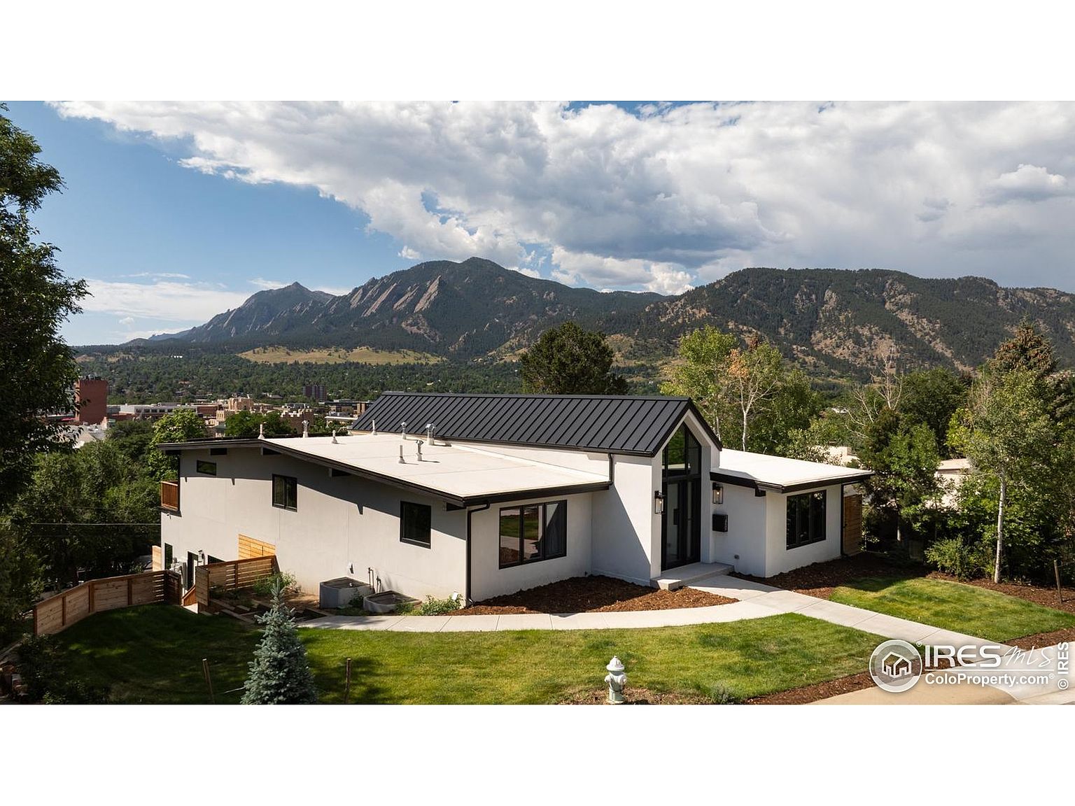 This is a front exterior view of a modern, single-family home with a white facade and a dark metal roof. The house features clean lines, large windows with dark frames, and a well-manicured lawn. In the background, there are mountains and trees, adding to the property's appeal and highlighting its location.
