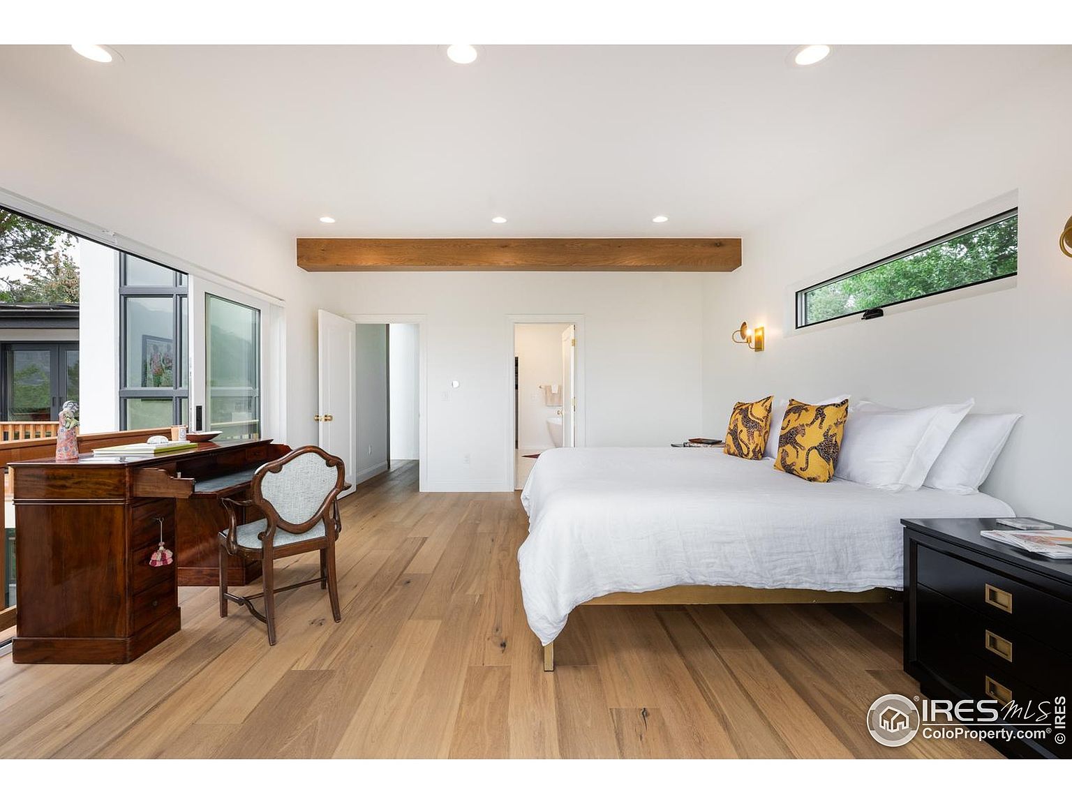 This is a bright and airy primary bedroom featuring hardwood floors, white walls, and a large window offering natural light. The room includes a bed with white linens and decorative pillows, a dark wood nightstand, and a vintage-style desk with a chair. A wooden beam adds a touch of rustic charm to the modern space.