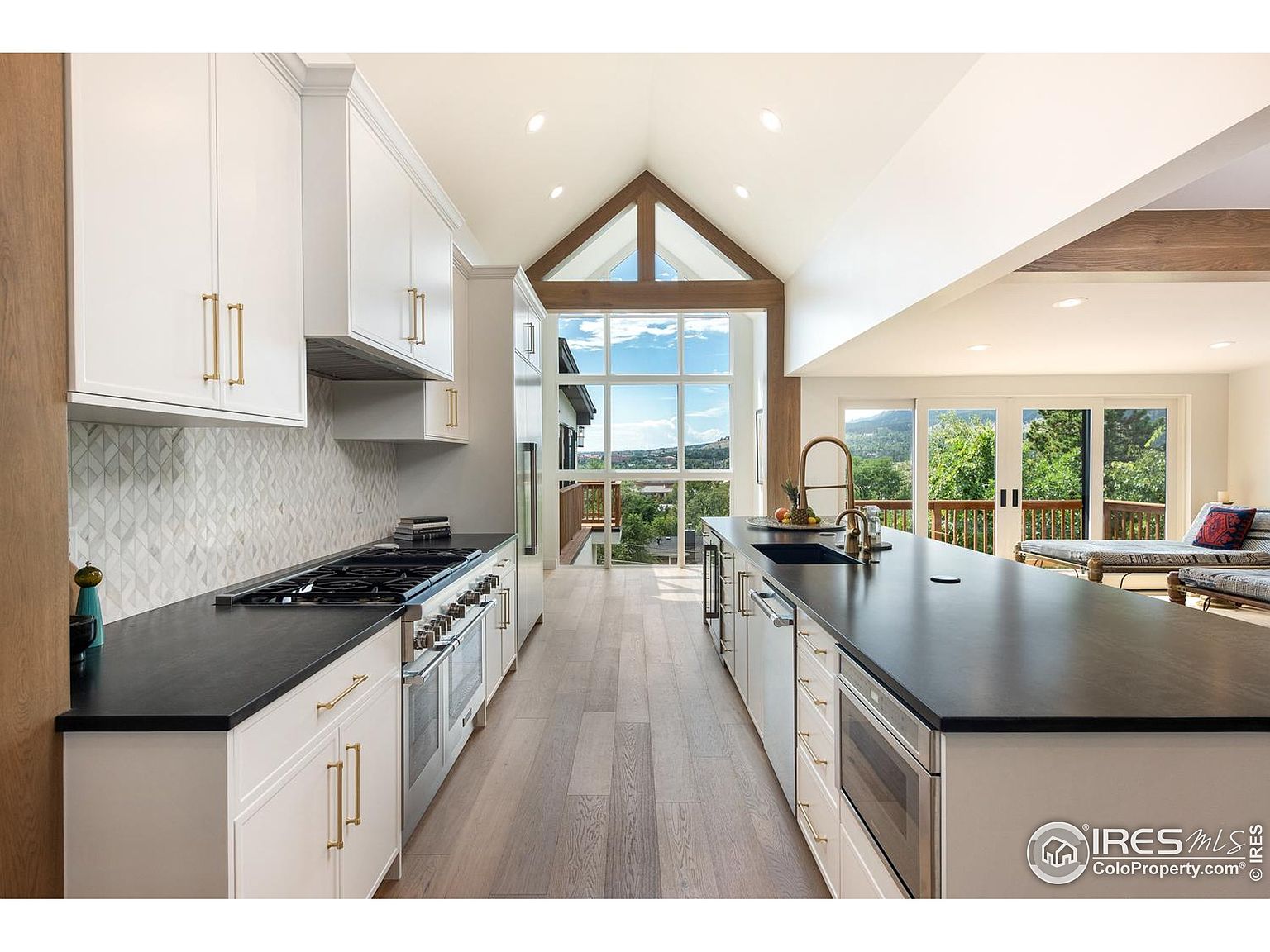 This is a bright and modern kitchen featuring white cabinetry with gold hardware, black countertops, and stainless steel appliances. The kitchen boasts a large island with a sink and a view of the outdoors through a large window. The open floor plan leads to a living area with sliding glass doors to a deck, creating a seamless indoor-outdoor living experience.