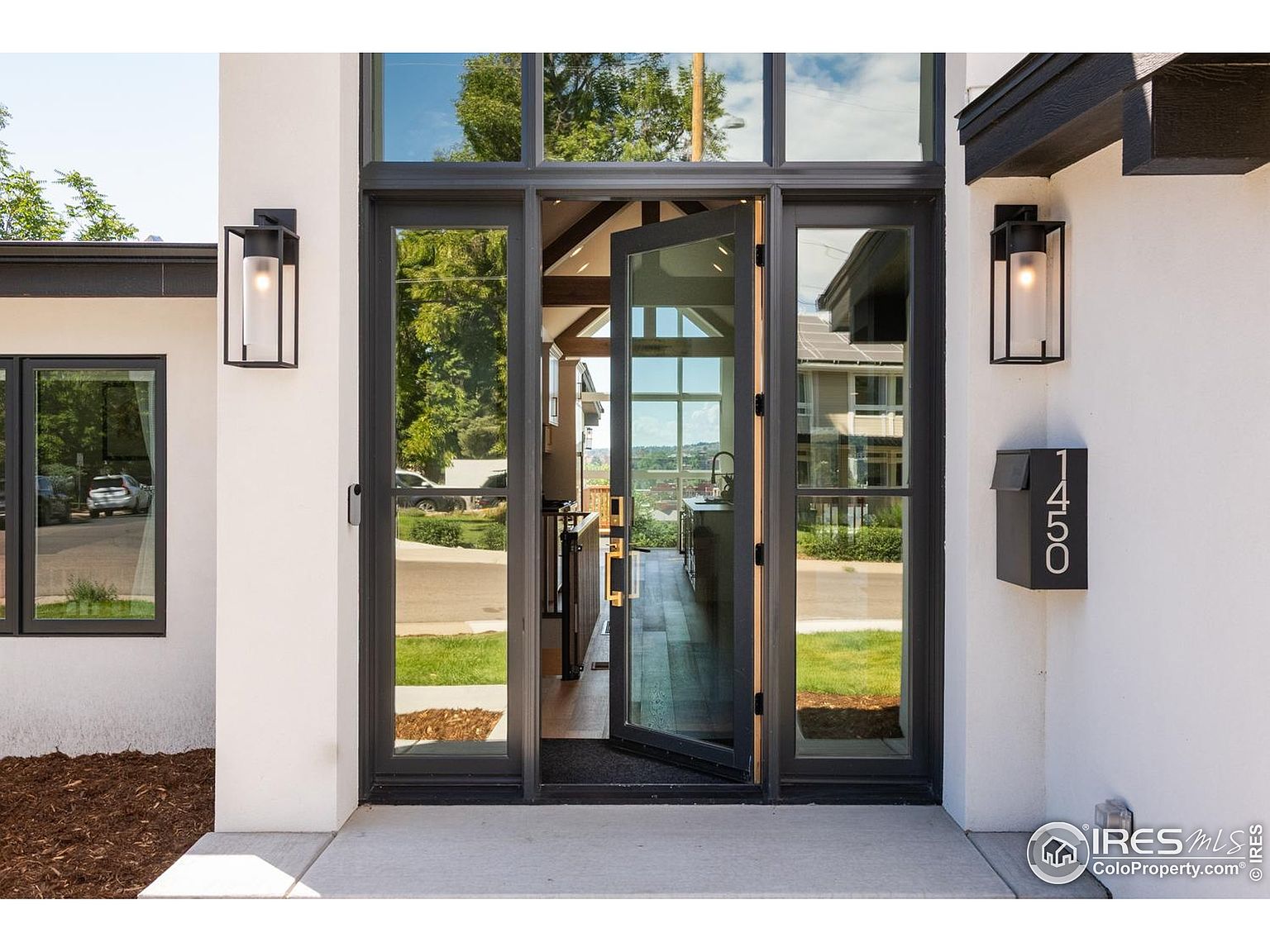 This image showcases a modern home's entryway, featuring a striking black-framed glass door and sidelights that flood the interior with natural light. The exterior is painted in a clean white, complemented by black accents on the roofline and light fixtures. A sleek mailbox with house numbers adds a contemporary touch, while the open door offers a glimpse into the home's stylish interior.