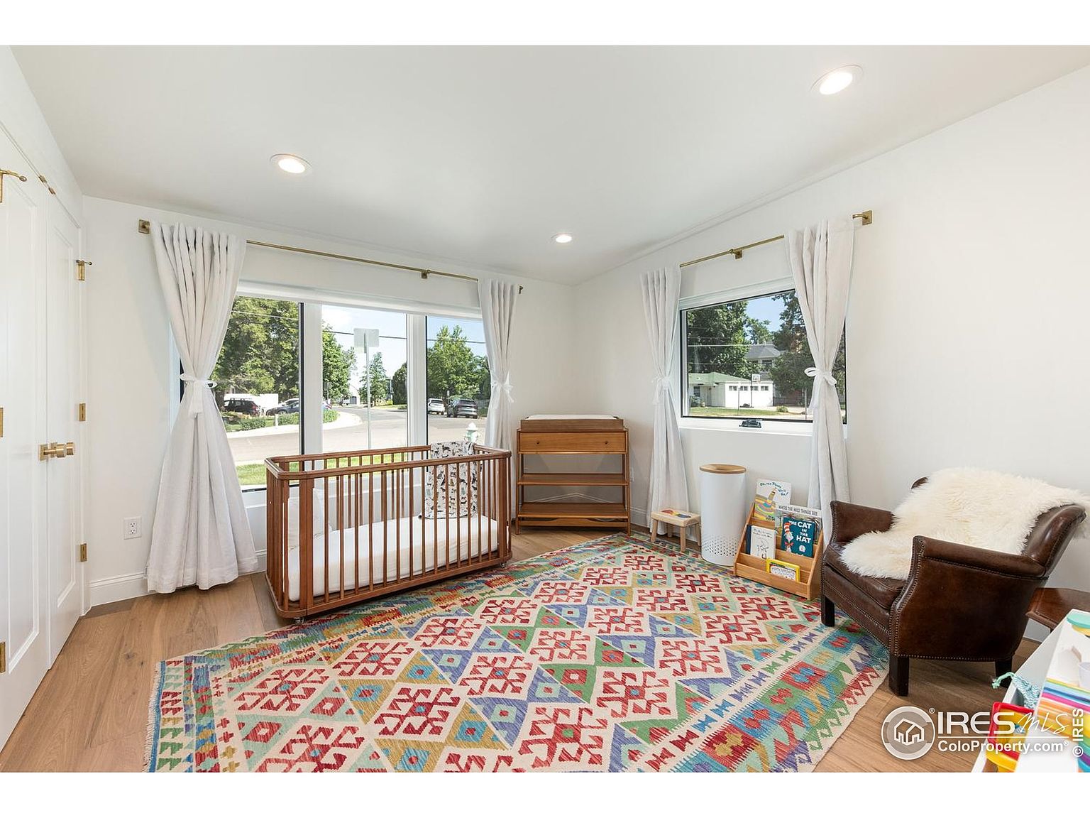 This is a bright and airy nursery featuring a wooden crib with white bedding, a colorful patterned rug, and a comfortable armchair with a fluffy throw. Natural light floods the room through two large windows, complemented by white walls and curtains, creating a serene and inviting space. A changing table and bookshelf add functionality to the room.