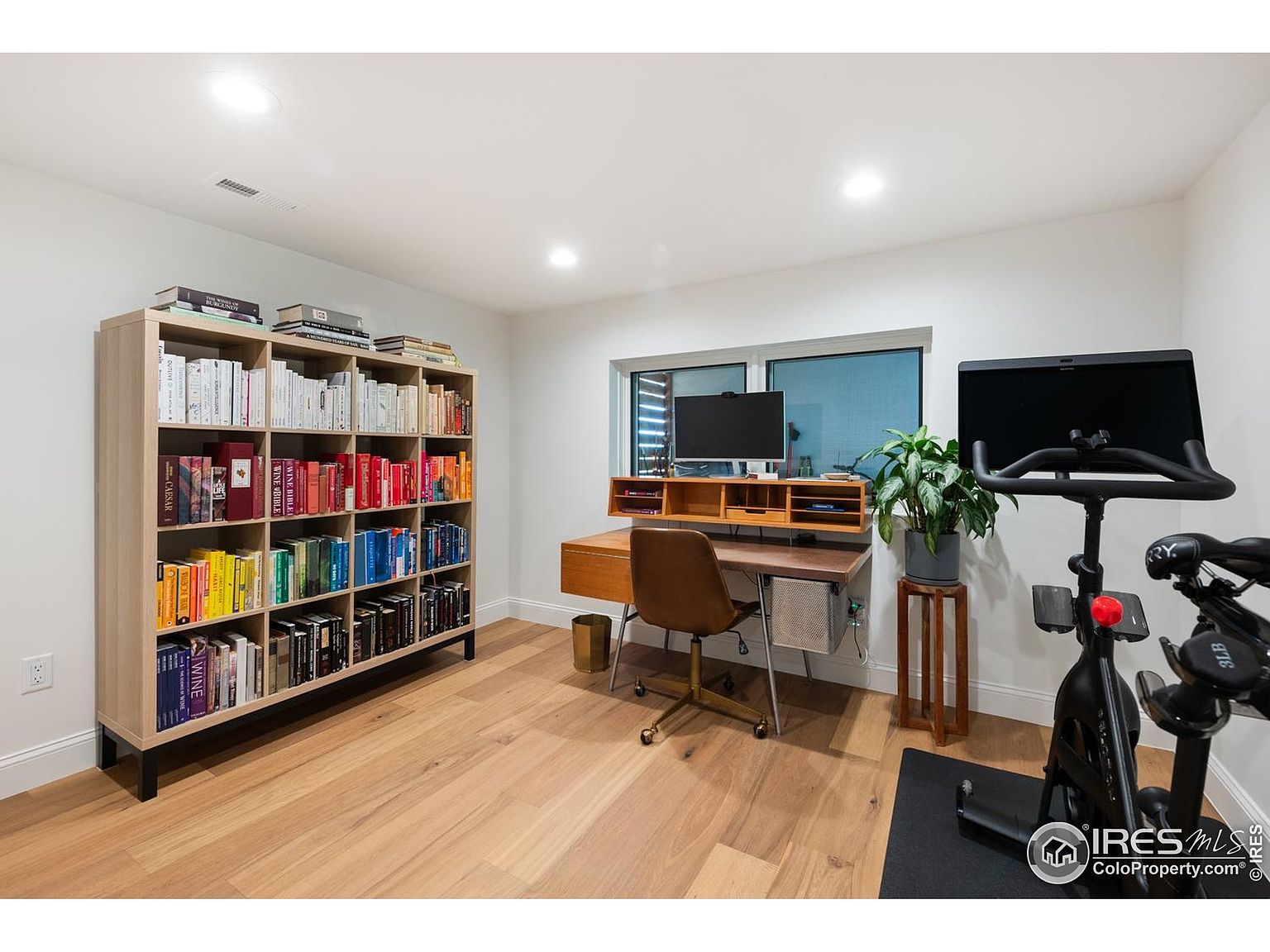 This interior shot showcases a well-organized home office space. A large bookshelf filled with books anchors one side of the room, while a desk with a computer and a plant sits near a window. An exercise bike is positioned on the right, suggesting a dual-purpose space for work and fitness.