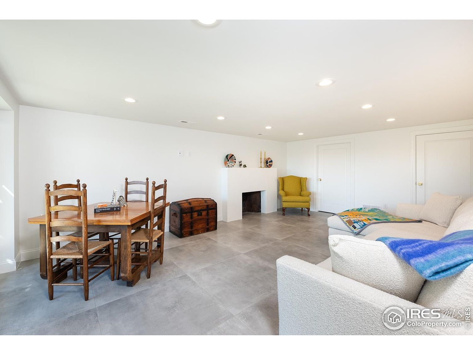 This interior shot showcases a spacious living area with a blend of rustic and modern elements. The room features a dining table with wooden chairs, a decorative chest, a white fireplace, and a comfortable-looking sofa with throw blankets. The gray tile flooring and white walls create a bright and airy atmosphere, making it an inviting space for relaxation and entertainment.