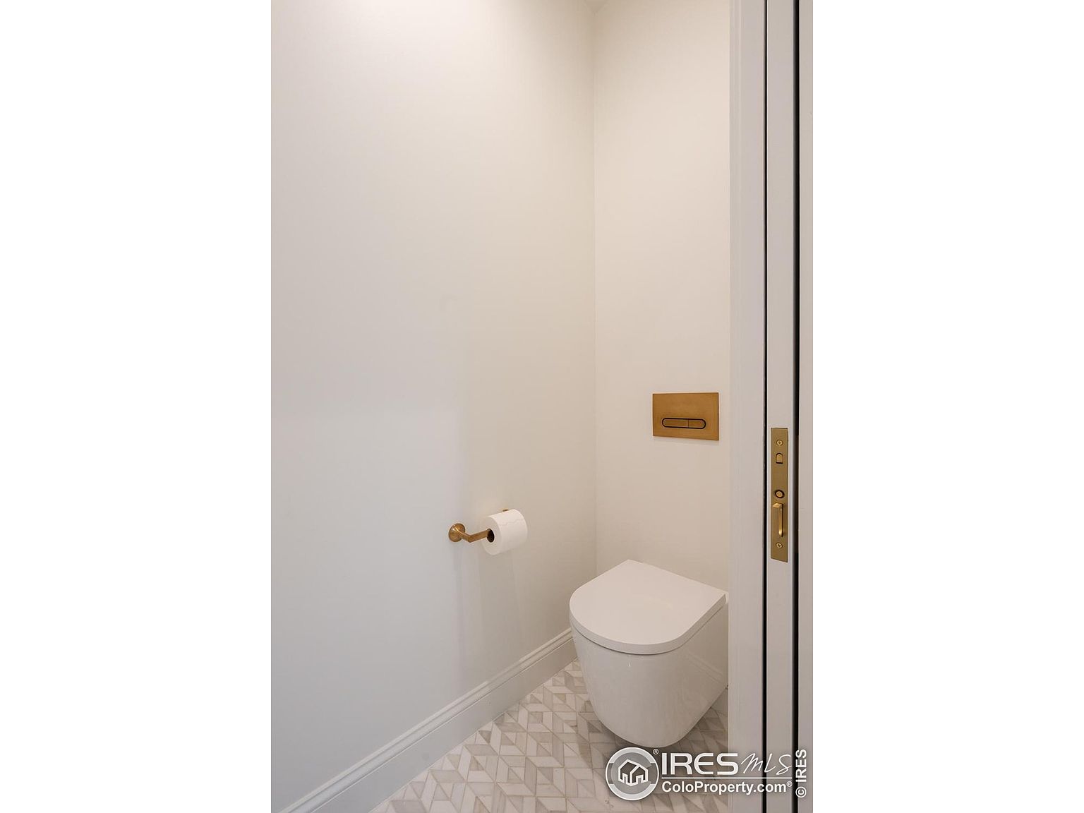 This is a well-lit, modern guest bathroom featuring a wall-mounted toilet with a sleek, minimalist design. The walls are painted in a neutral tone, complemented by a geometric patterned tile floor. A gold toilet paper holder and flush plate add a touch of elegance, while the white door frame provides a clean contrast.