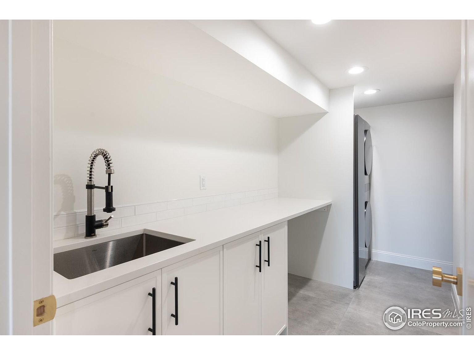 This is a bright and clean laundry room featuring white cabinetry with sleek black hardware, a stainless steel sink with a modern faucet, and a light gray tile floor. A stacked washer and dryer unit is visible in the background, and the room is well-lit with recessed lighting, creating a functional and aesthetically pleasing space.