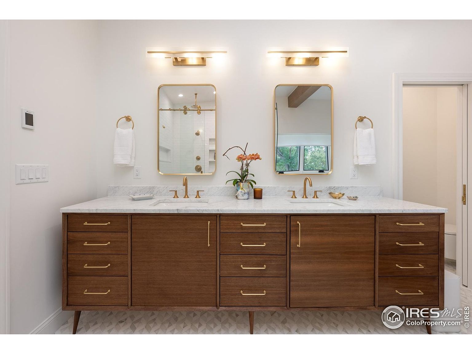 This is a well-lit primary bathroom featuring a double vanity with a white marble countertop and dark wood cabinetry. Two gold-framed mirrors hang above each sink, complemented by matching gold light fixtures and towel rings. The flooring is a patterned tile, and a glimpse of the shower and toilet area can be seen through the doorway, creating a luxurious and modern feel.
