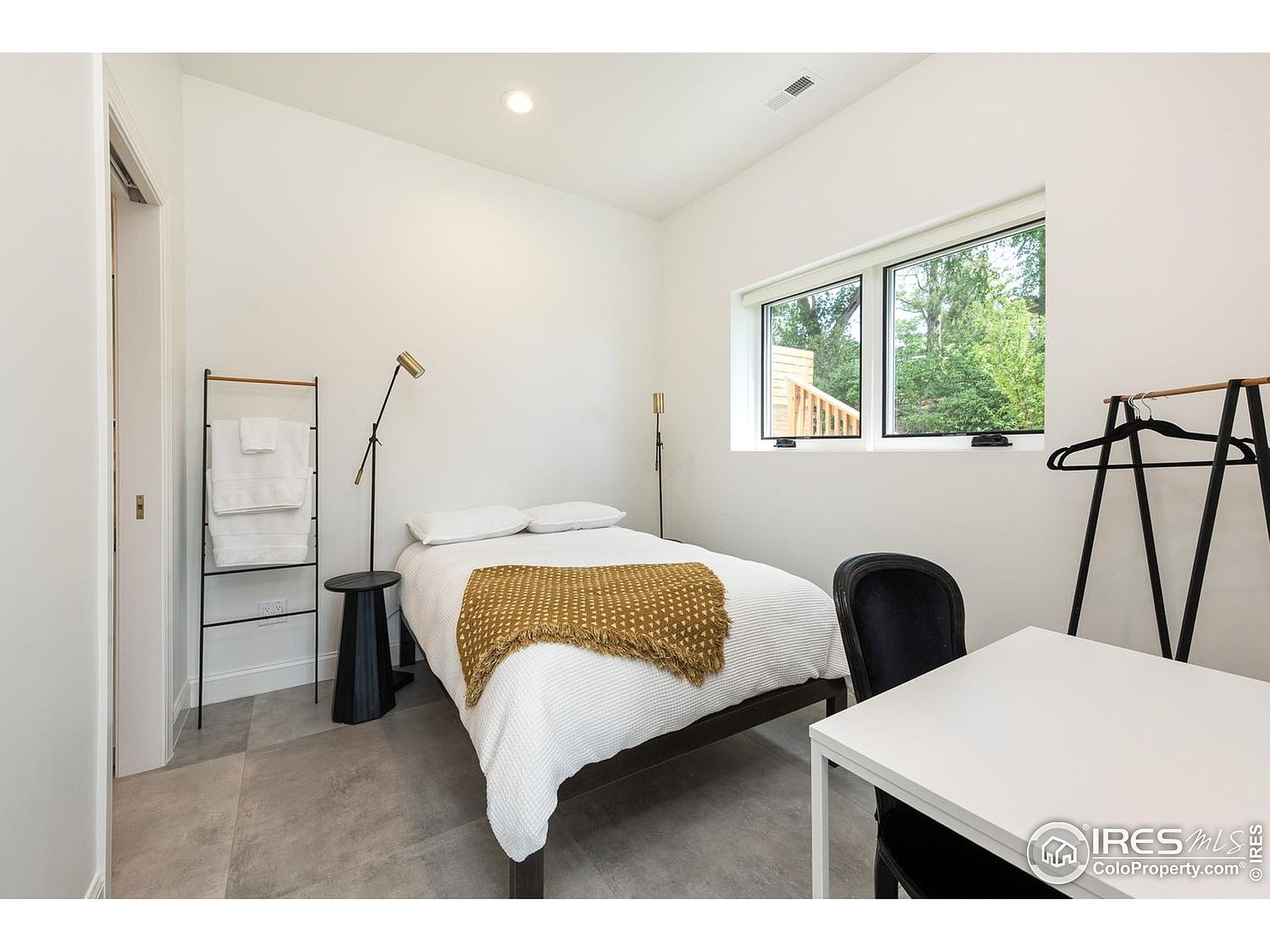 This is a well-lit guest bedroom featuring a simple yet stylish design. The room includes a bed with white linens and a mustard-colored throw, a small side table, a modern floor lamp, and a desk area with a black chair. A window provides natural light and a view of the outdoors, while a towel rack and clothes rack add functional elements.