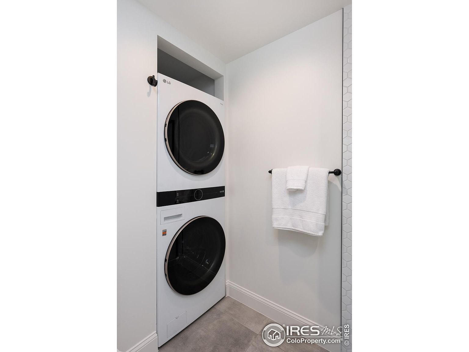 This is a well-organized laundry room featuring a stacked LG washer and dryer in white. A towel rack with neatly folded white towels is mounted on the adjacent wall. The room has a clean and modern aesthetic with light gray flooring and white walls, creating a functional and visually appealing space.