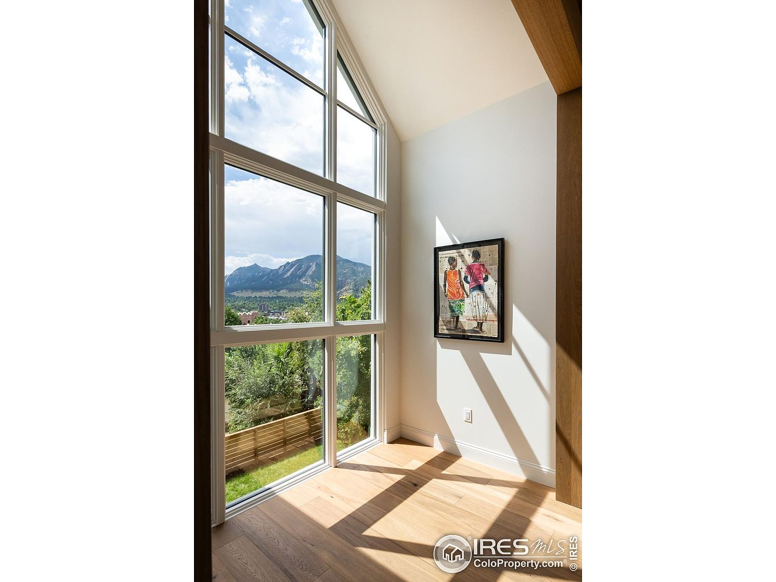 This sunroom features a large, multi-pane window offering a scenic view of mountains and greenery. The room has light hardwood flooring and a minimalist design, with a framed artwork on the wall. Natural light floods the space, creating a bright and airy atmosphere.
