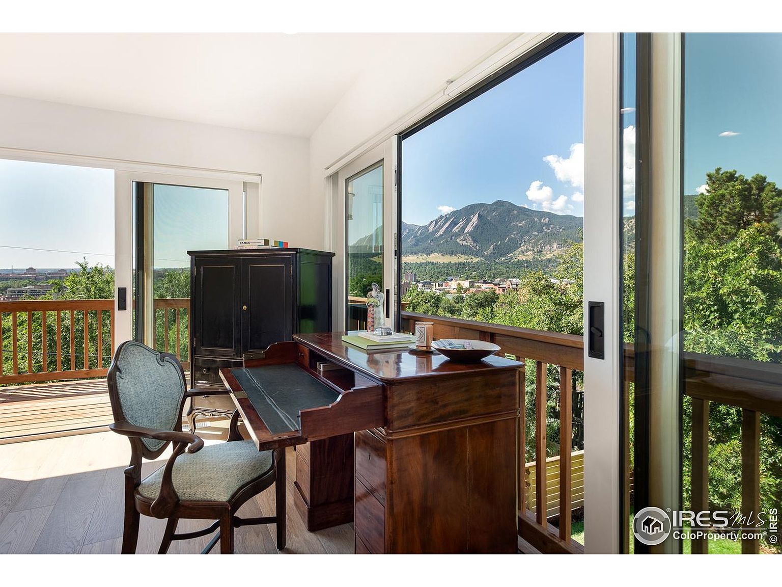 This is an interior shot of a home office or study area. The room features a wooden desk with a leather writing surface, a vintage-style chair, and a dark cabinet. Large sliding glass doors open onto a balcony with a view of mountains and trees, creating a bright and airy workspace.