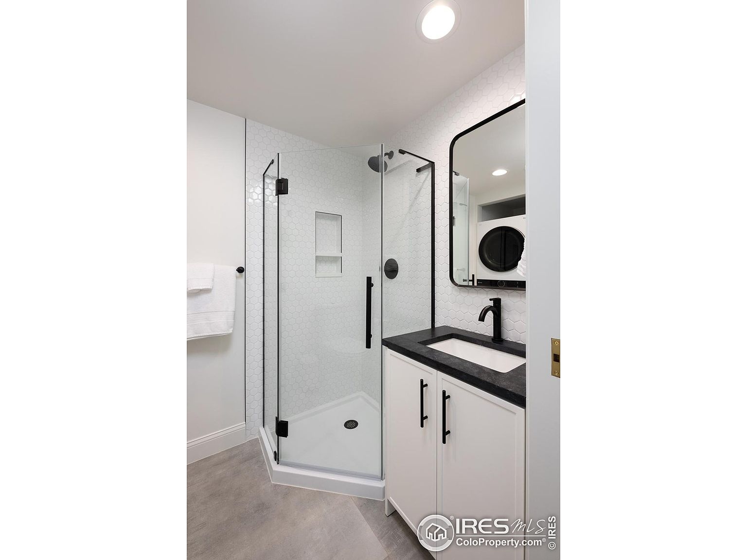 This is a modern guest bathroom featuring a glass-enclosed shower with white hexagon tile walls and a built-in niche. The vanity has a black countertop, white cabinets, and black hardware, complemented by a black-framed mirror. The flooring is a light gray, and the overall design is clean and contemporary.