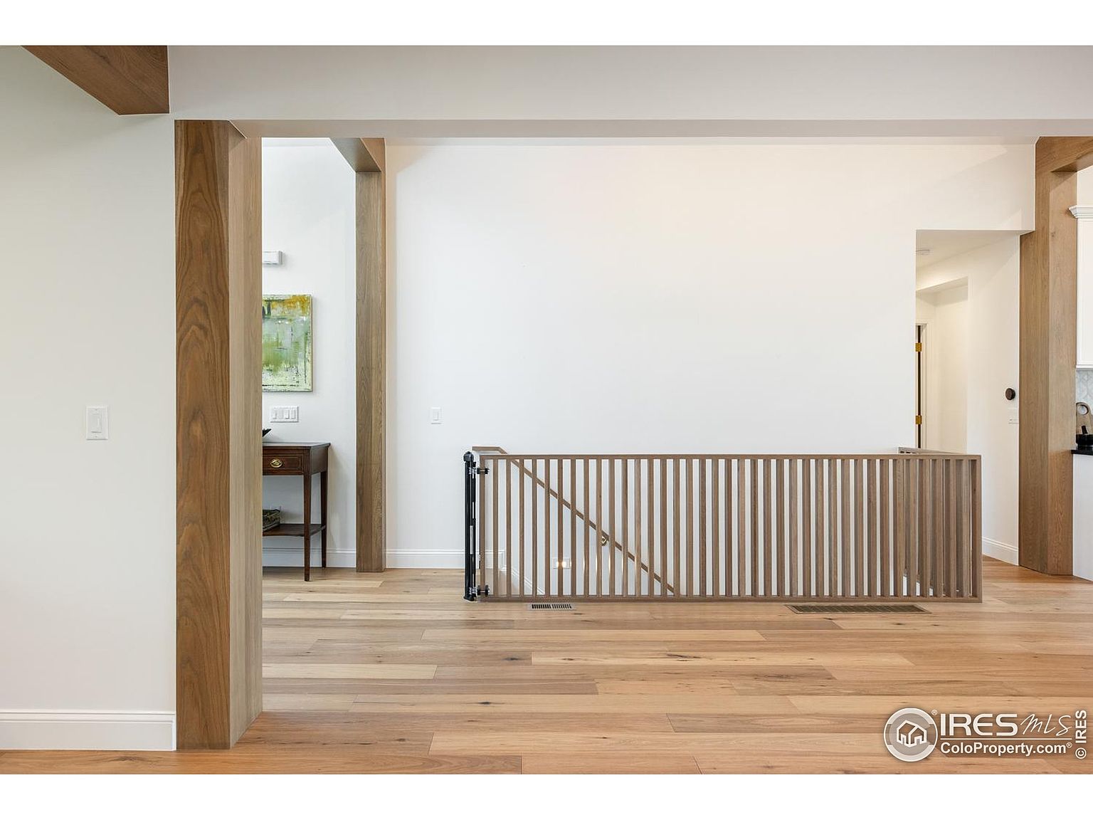 This interior shot showcases a bright and open hallway with hardwood flooring and wooden support beams. A wooden railing acts as a barrier, and a small table with a painting above it is visible in the background. The space is well-lit and appears to be part of a modern or updated home.