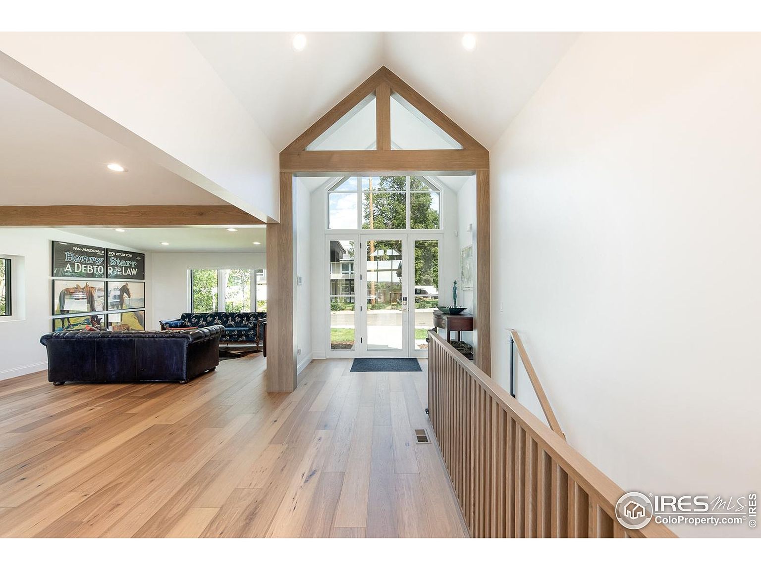 This interior shot showcases a bright and spacious hallway with light wood flooring and white walls. A striking architectural feature is the wood-framed entryway with large windows, allowing natural light to flood the space. A wooden railing lines the staircase, adding warmth and texture to the modern design.