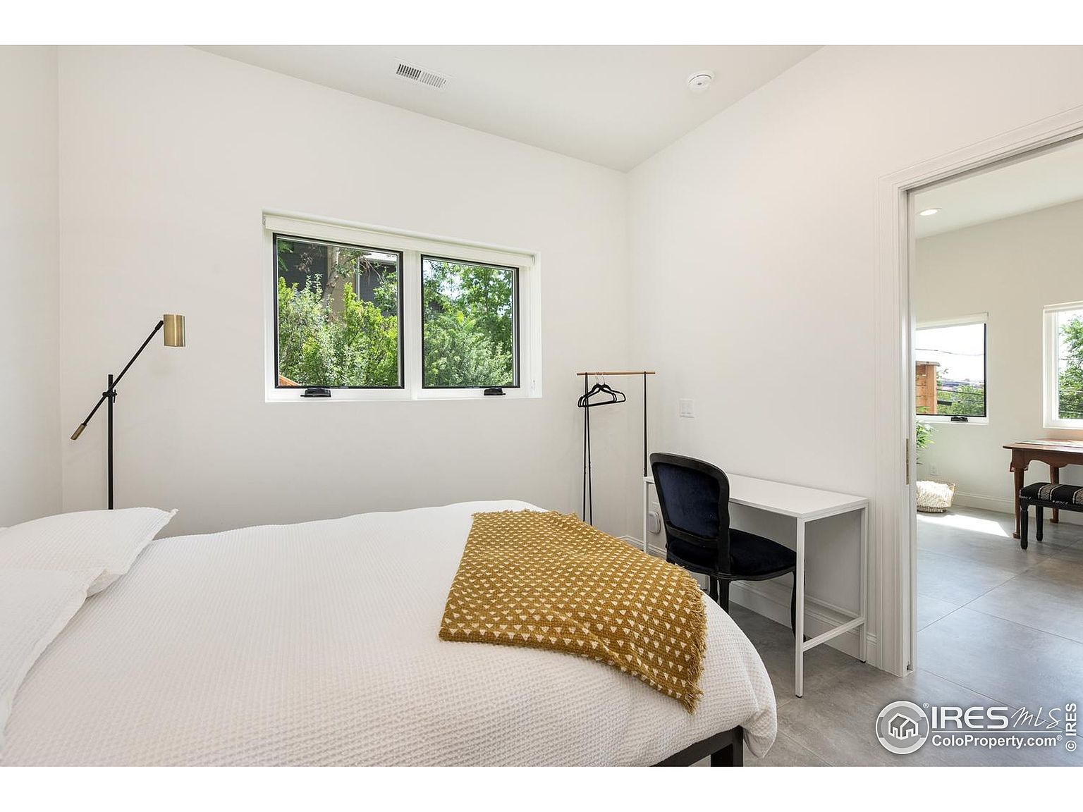 This is a bright and minimalist bedroom featuring a bed with white linens and a mustard-colored throw, a desk with a blue chair, and a clothing rack. A window provides natural light and a view of greenery outside. The room has a clean and modern aesthetic, suitable for a guest or child's bedroom.