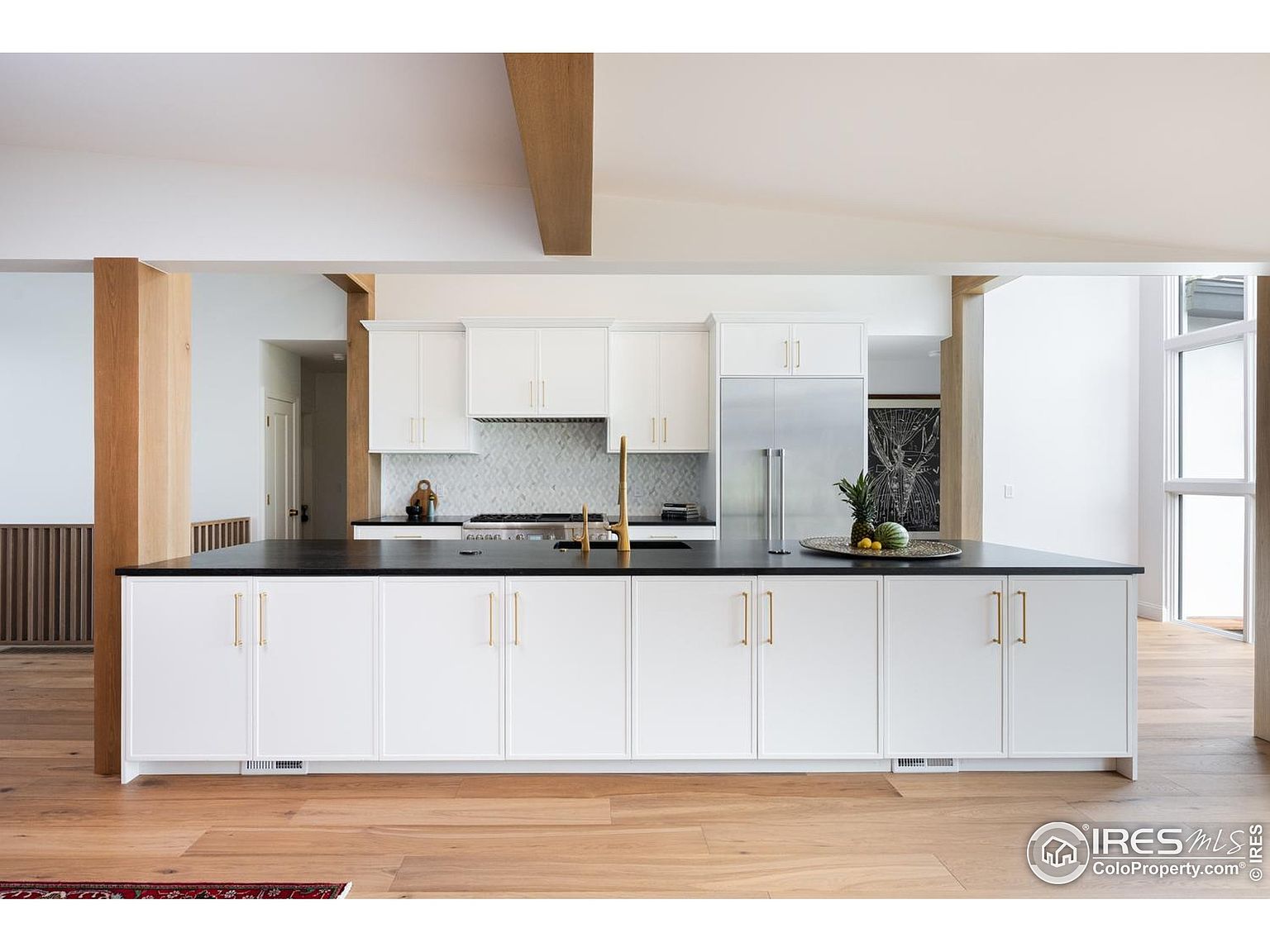 This is a bright and modern kitchen featuring a large island with a black countertop and white cabinetry with gold hardware. Stainless steel appliances are integrated into the design, and the space is open with wooden beams and flooring. The kitchen exudes a clean and sophisticated aesthetic.