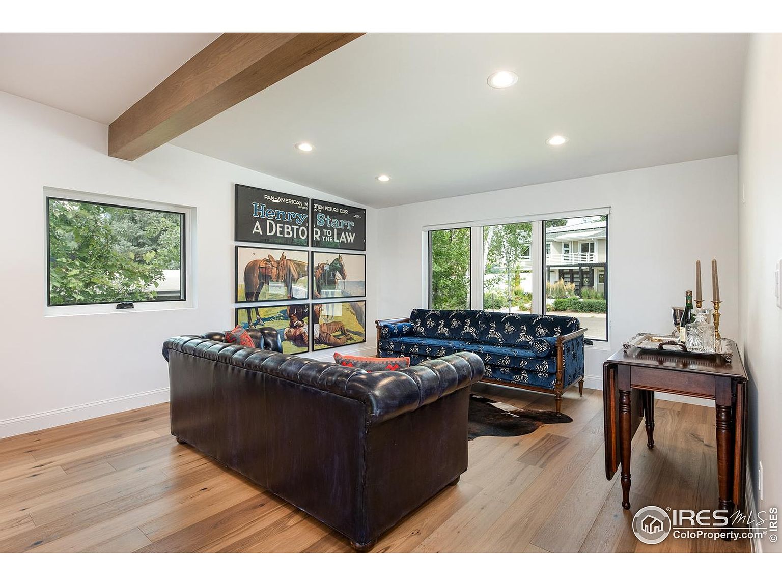 This is an interior shot of a living room featuring hardwood floors, white walls, and a wooden beam on the ceiling. The room is furnished with a dark leather sofa, a blue patterned sofa, and a dark wood side table. Artwork adorns the wall, and natural light streams in through the windows, creating a bright and inviting space.