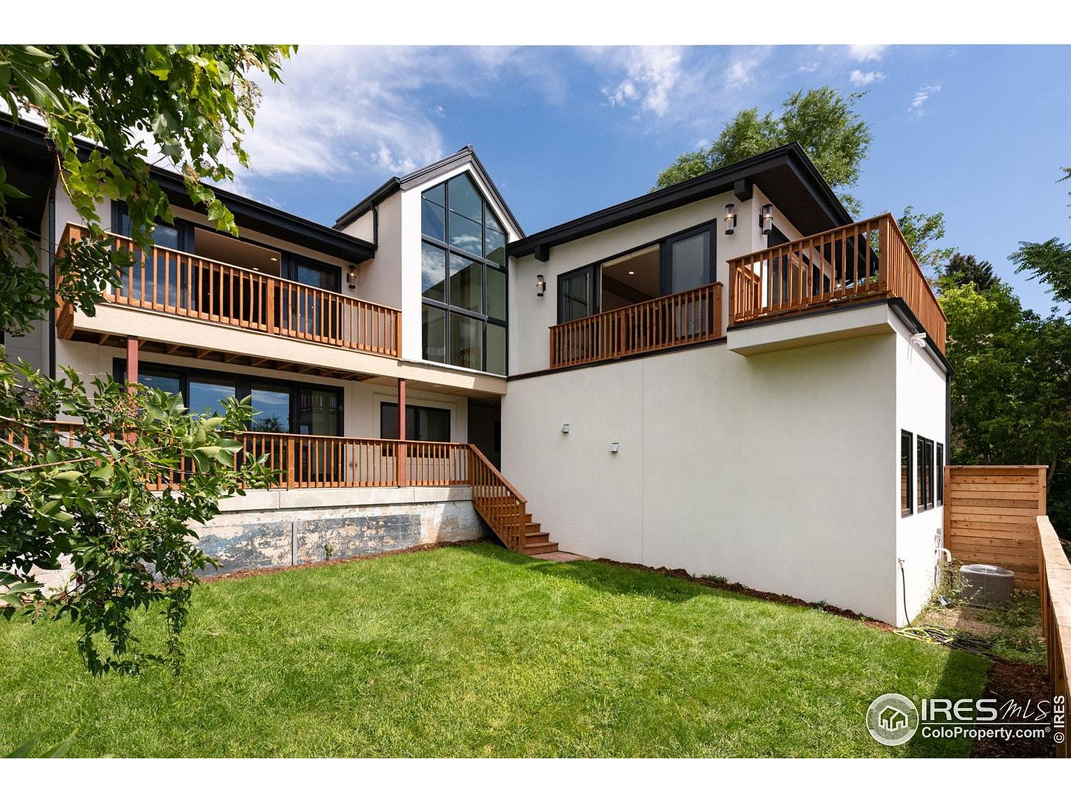 This is a rear view of a modern two-story home with a well-maintained lawn. The house features multiple wooden balconies, a large window section, and a white exterior. The design emphasizes outdoor living and natural light, creating an inviting and stylish impression.