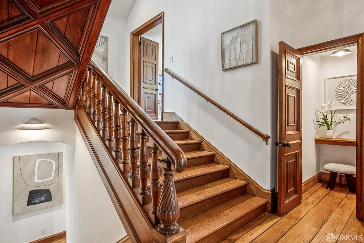 This interior view captures a classic wooden staircase with ornate balusters and a rich, warm-toned wood finish. To the right, an open doorway leads into a small, bright alcove featuring a floating shelf, a decorative shell art piece, and a small stool. The walls are painted a crisp white, accented by framed artwork and a unique wood-paneled ceiling detail above the landing, creating a sophisticated and traditional atmosphere.